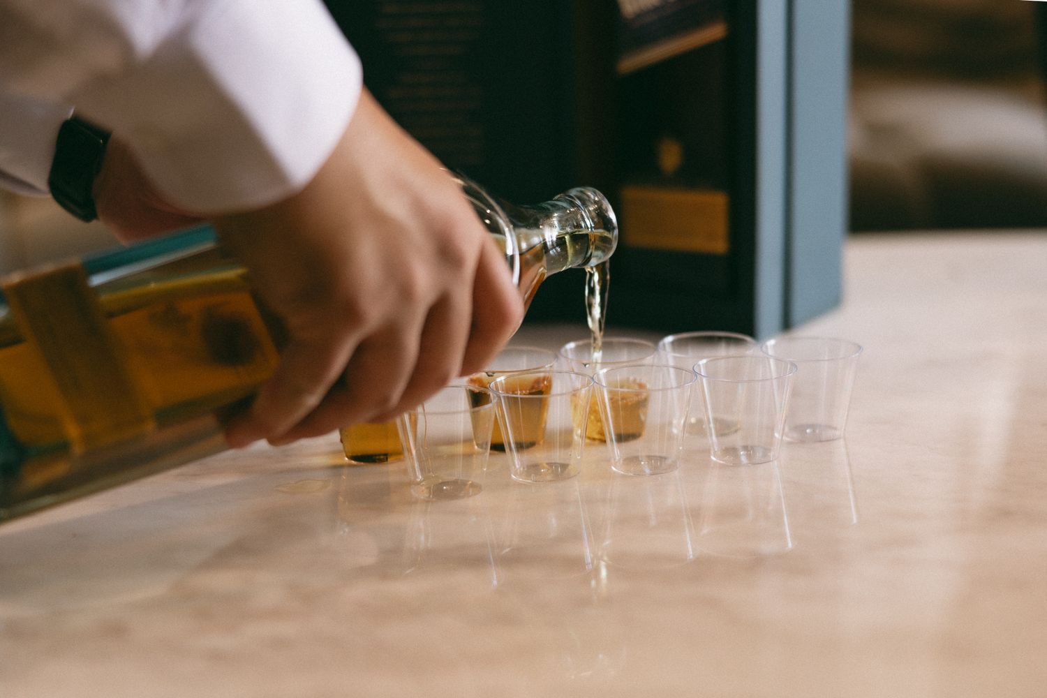 Bartender pouring whiskey from a bottle into a row of shot glasses on a marble bar counter.