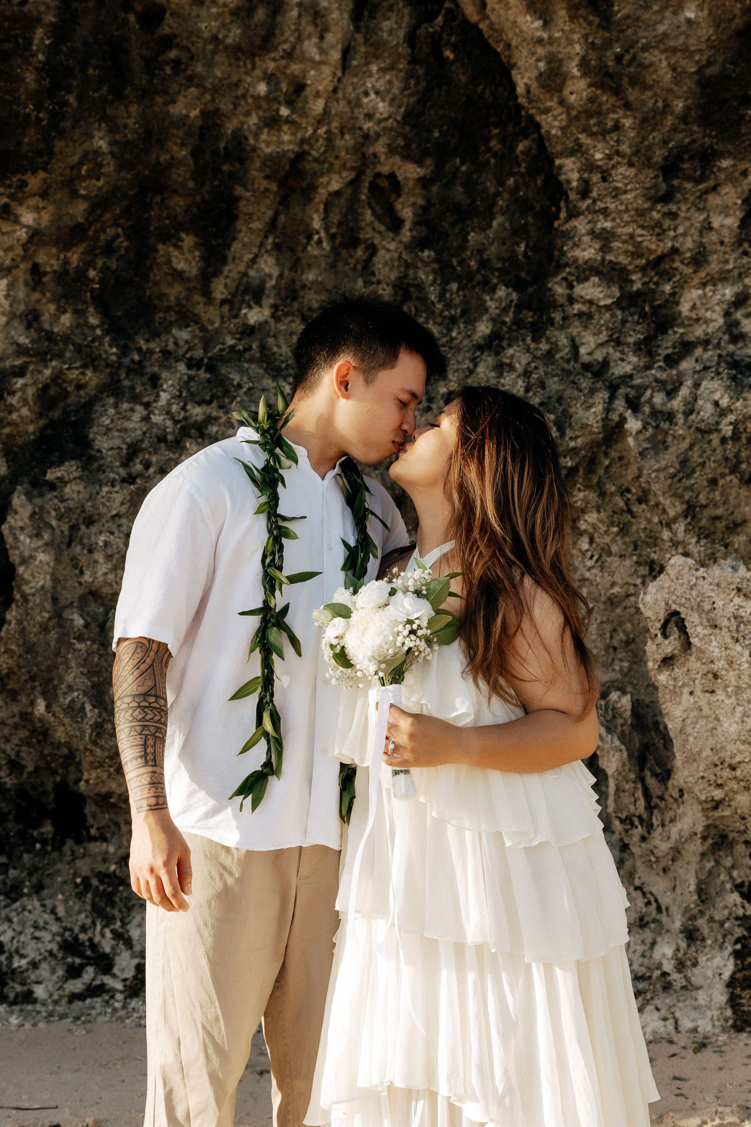 Couple sharing a kiss on a beach wedding, groom in white shirt with lei, bride in white dress holding bouquet.
