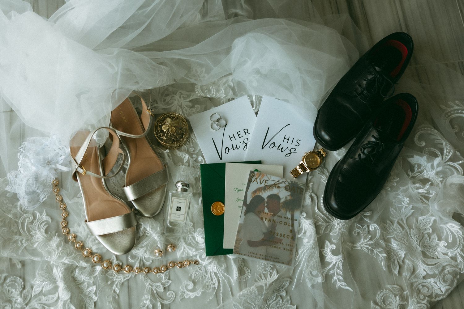 Wedding flat lay featuring bridal heels, groom dress shoes, vow books, pearl necklace, and Jo Malone perfume on lace gown.