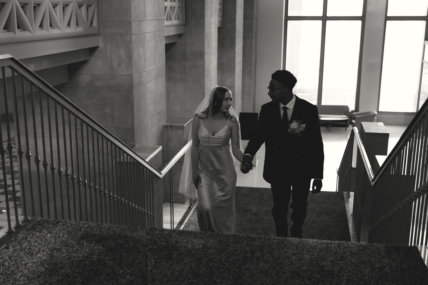 Black and white photo of a bride and groom holding hands while walking up elegant stairs at their wedding venue.