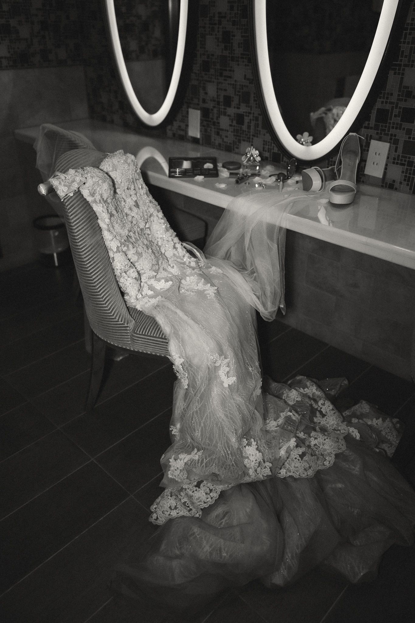 Black and white image of a lace wedding dress draped over a chair near a vanity mirror.