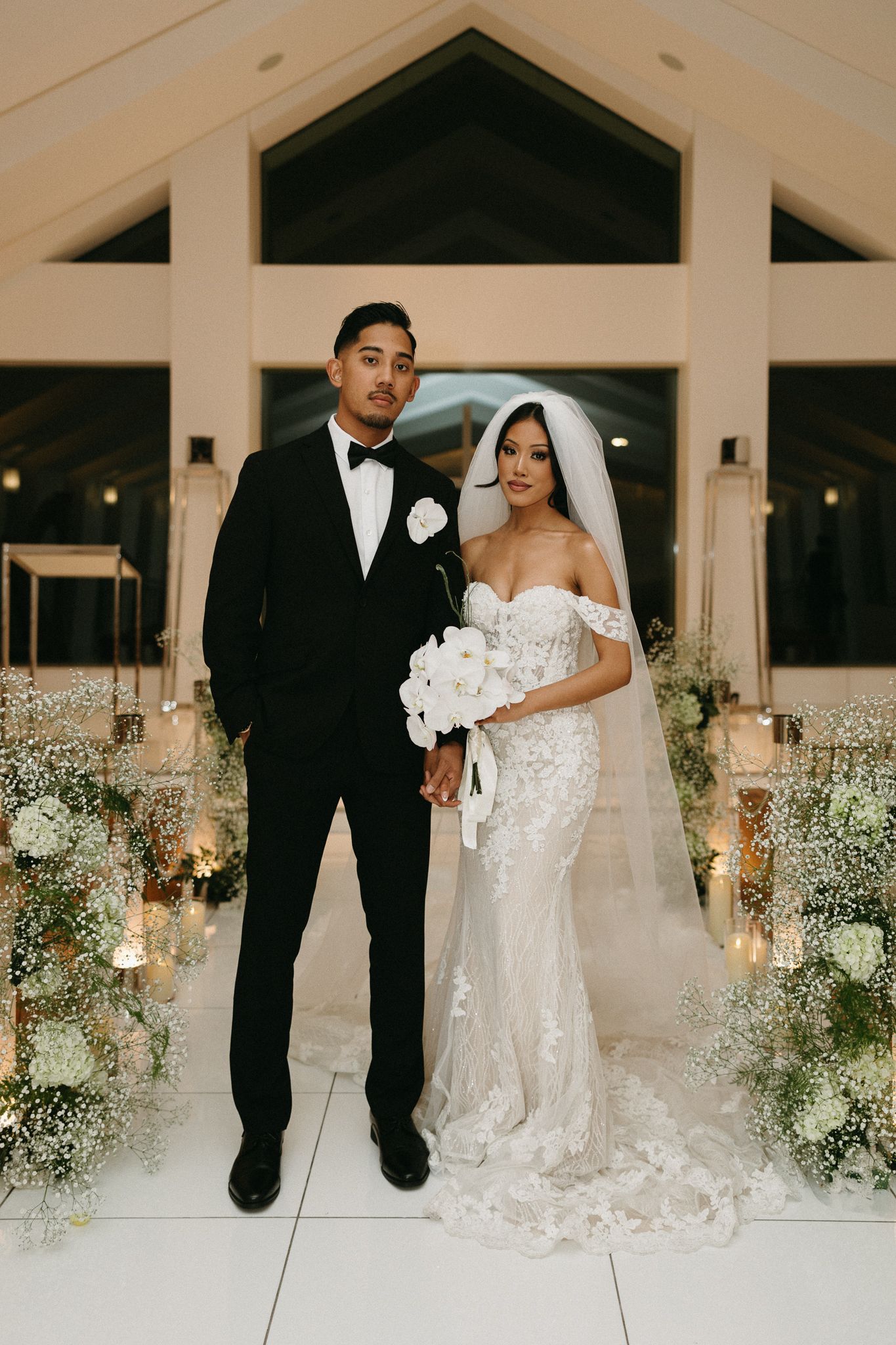 Bride and groom pose at elegant indoor wedding ceremony with white floral arrangements and candlelight.
