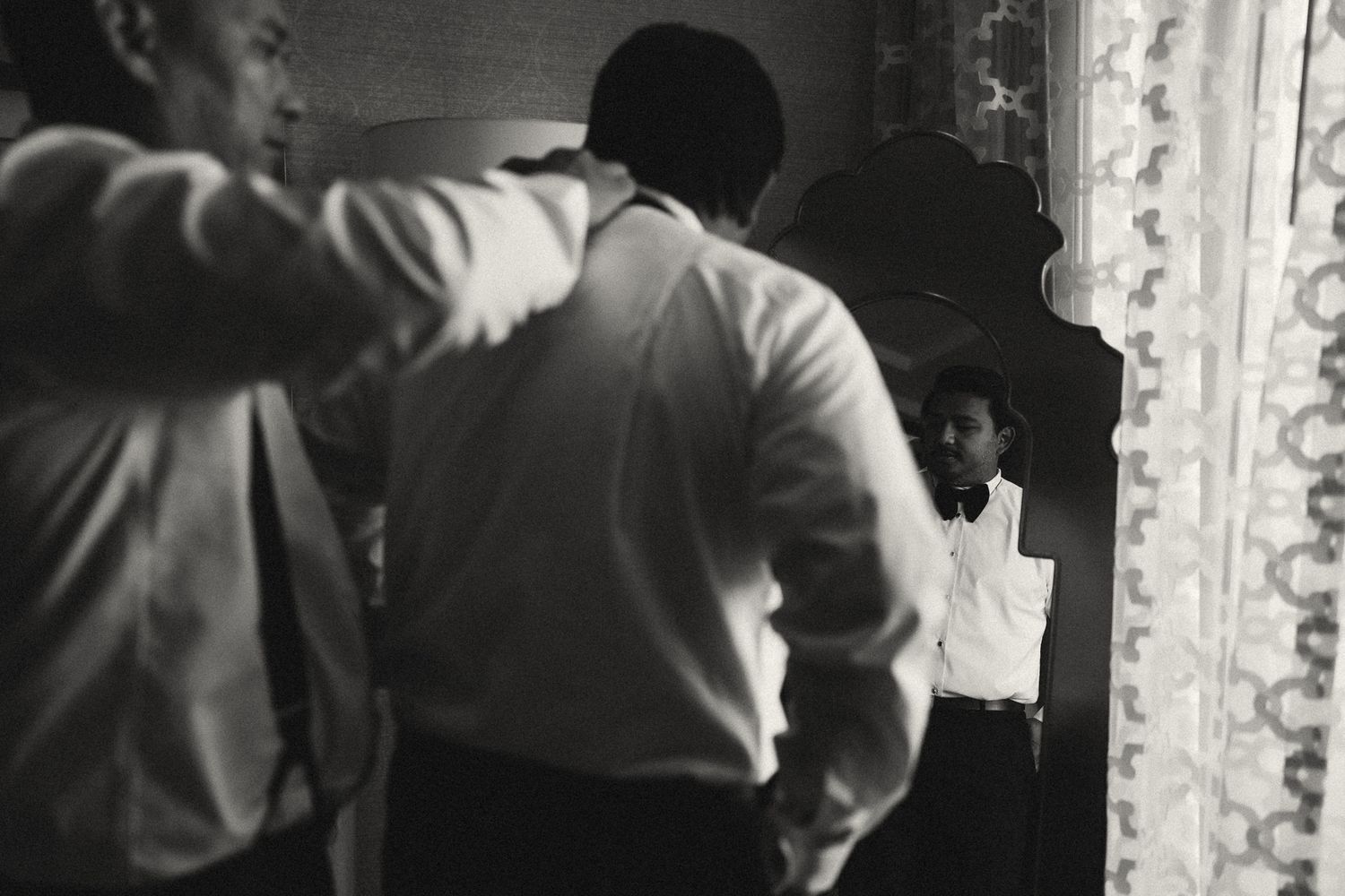 Black and white photo of two men in dress shirts preparing, with one adjusting his tie reflected in an ornate mirror.