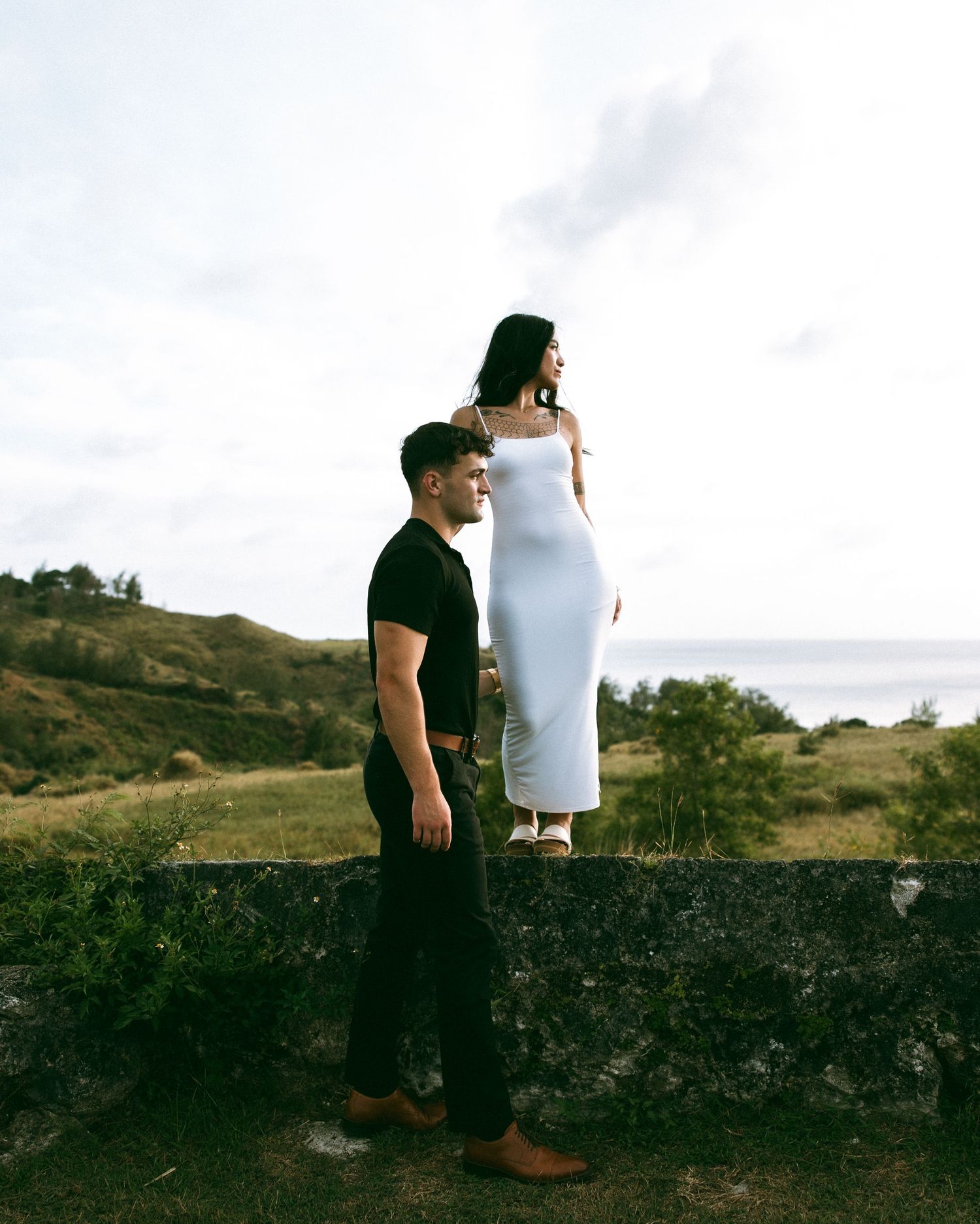 A pregnant woman in a white dress stands on a stone wall beside a man in black, with scenic hills behind them.