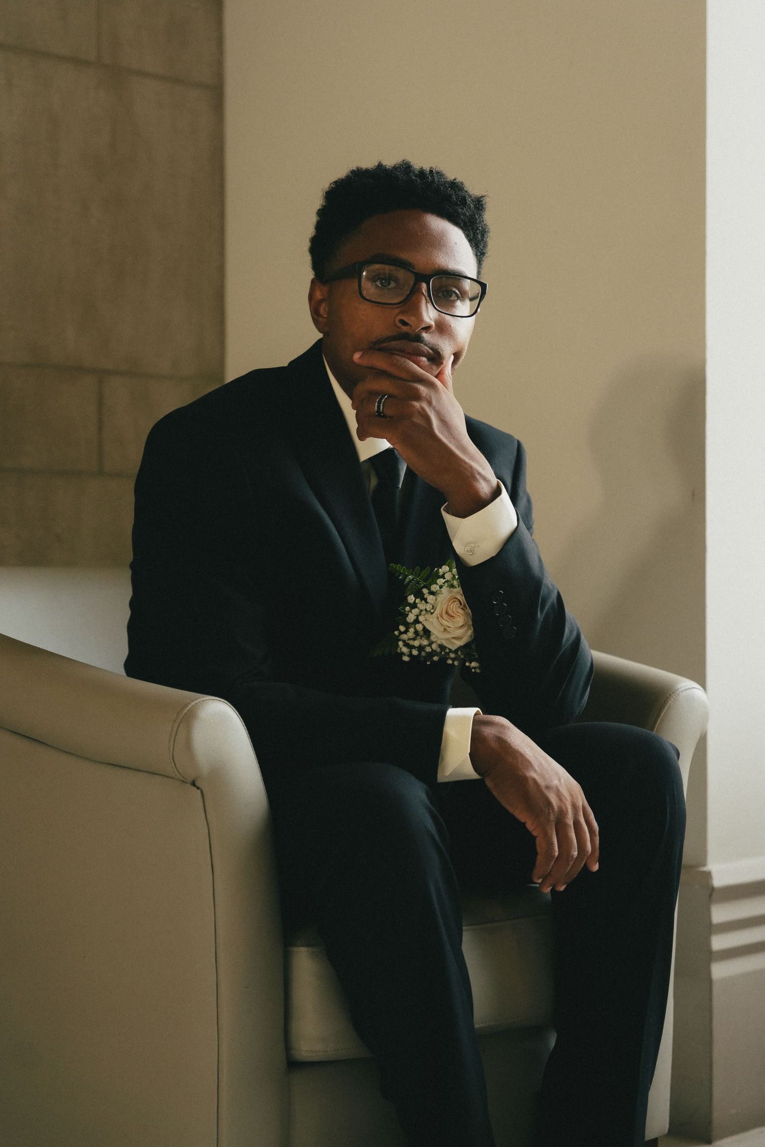 Groom in a black suit with glasses and boutonniere sitting thoughtfully in a chair at his wedding.