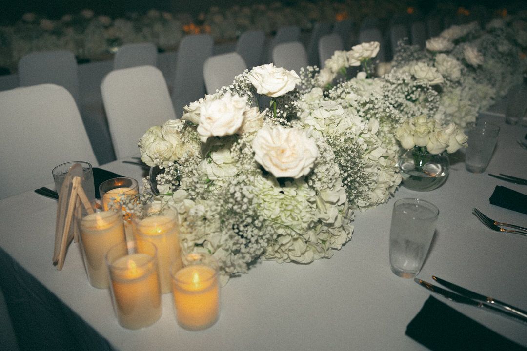 Elegant wedding table centerpiece with white roses, baby's breath, and glowing candles on a white linen tablecloth.