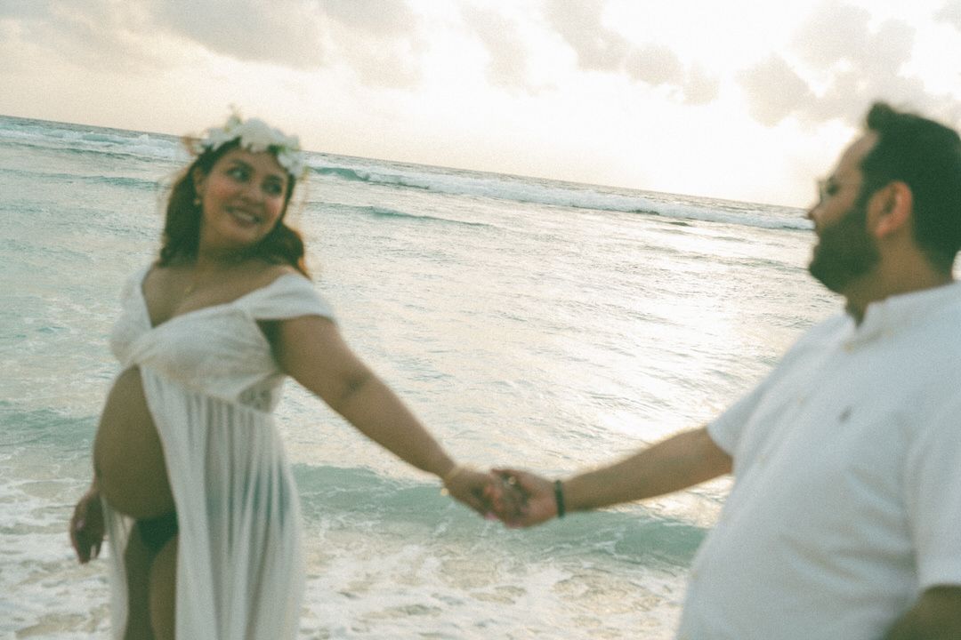Pregnant woman in white dress holds hands with man on tropical beach at sunset.