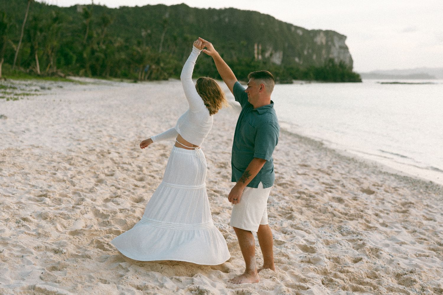 Couple dancing barefoot on a white sand beach, woman in flowing white dress, man in green shirt and white shorts.