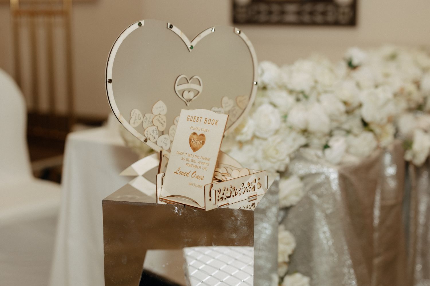Elegant wedding card box shaped like a heart, displayed on a silver stand with white floral arrangements in the background.