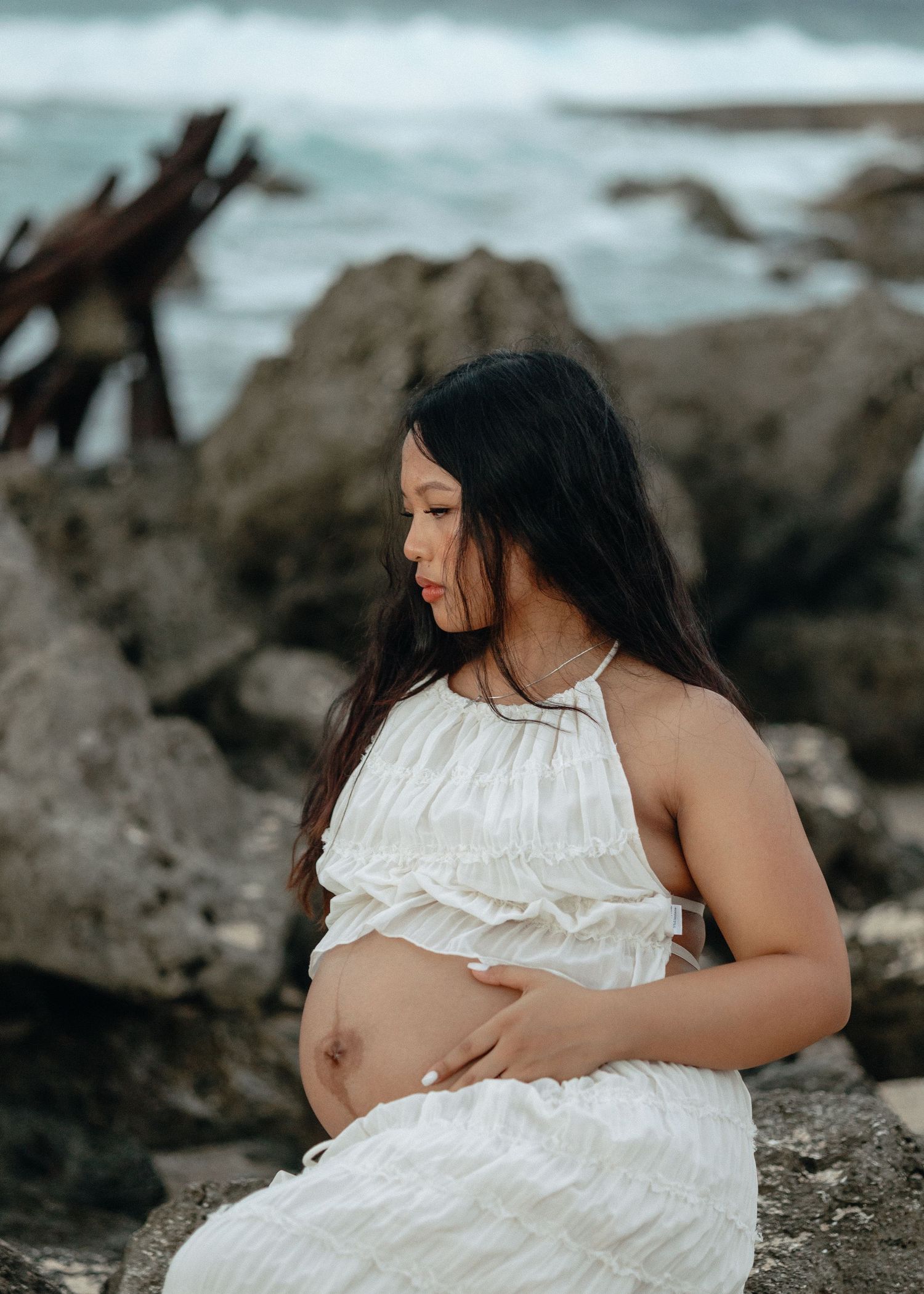 Pregnant woman in white dress cradling her baby bump on rocky beach with ocean waves in background.
