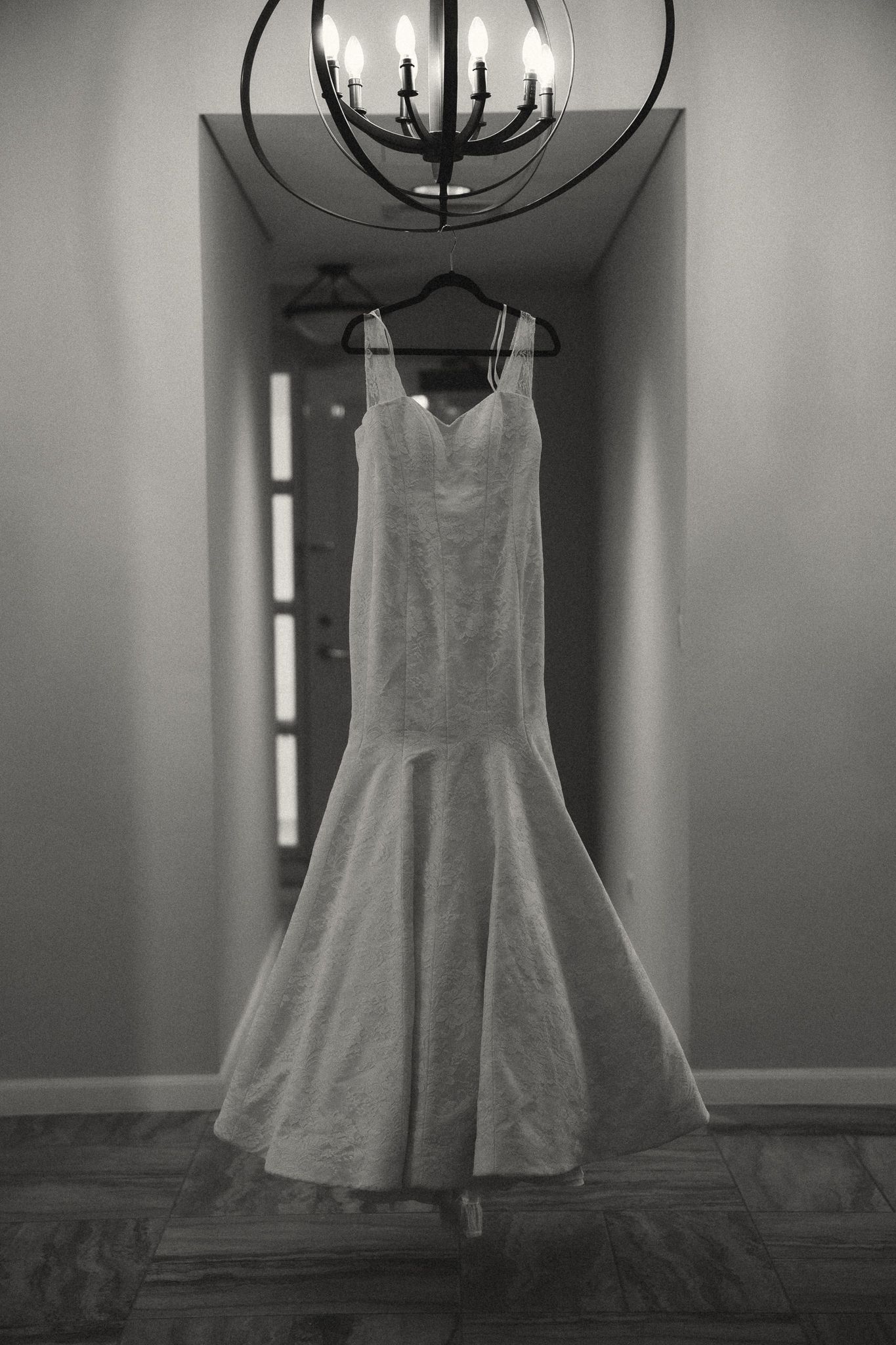 Black and white photo of an elegant mermaid wedding dress hanging from a chandelier in a hallway.