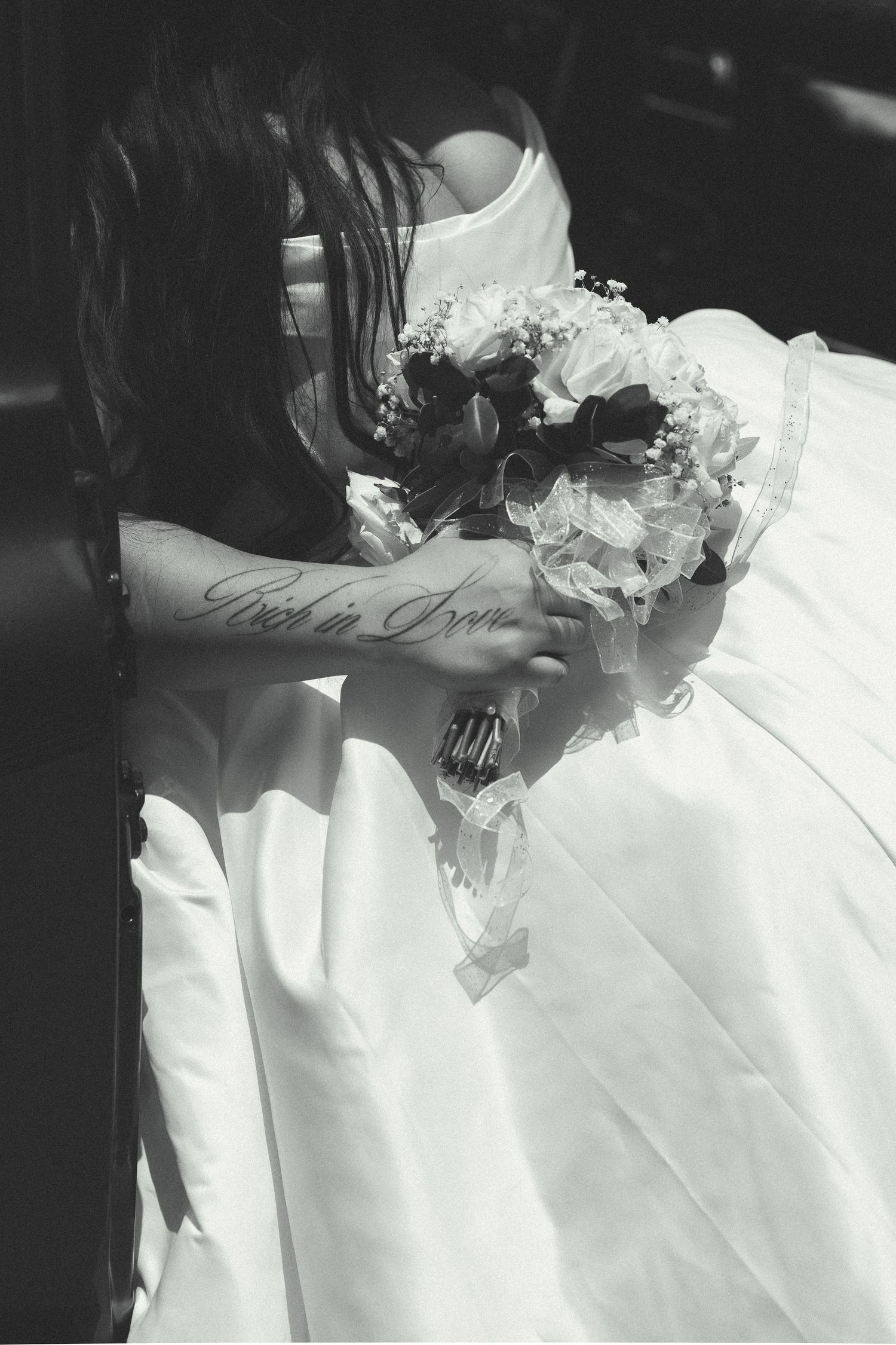 Black and white photo of bride in white gown holding floral bouquet with arm tattoo visible.