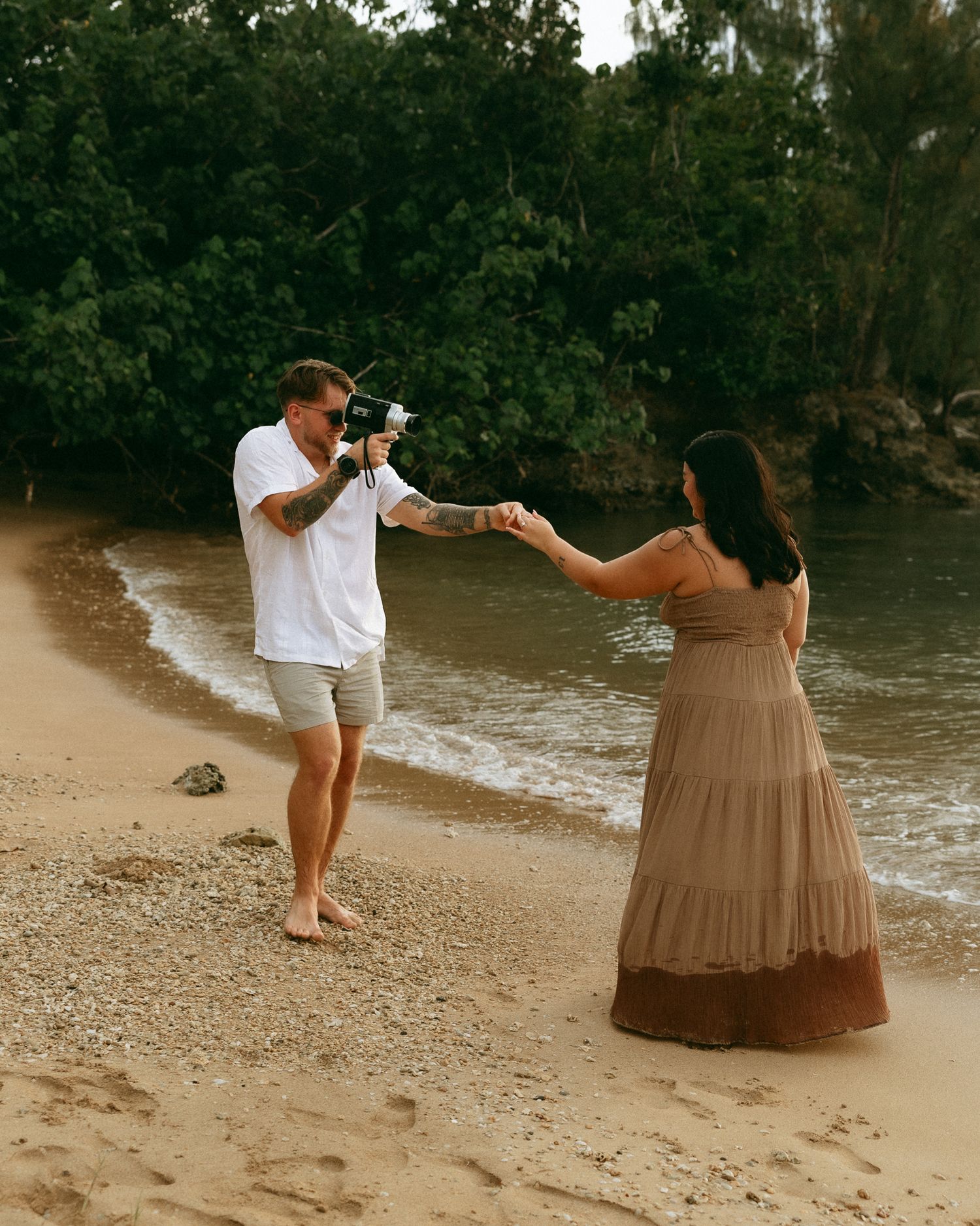 A man photographs a woman in a brown maxi dress on a tropical sandy beach, holding hands near the ocean.