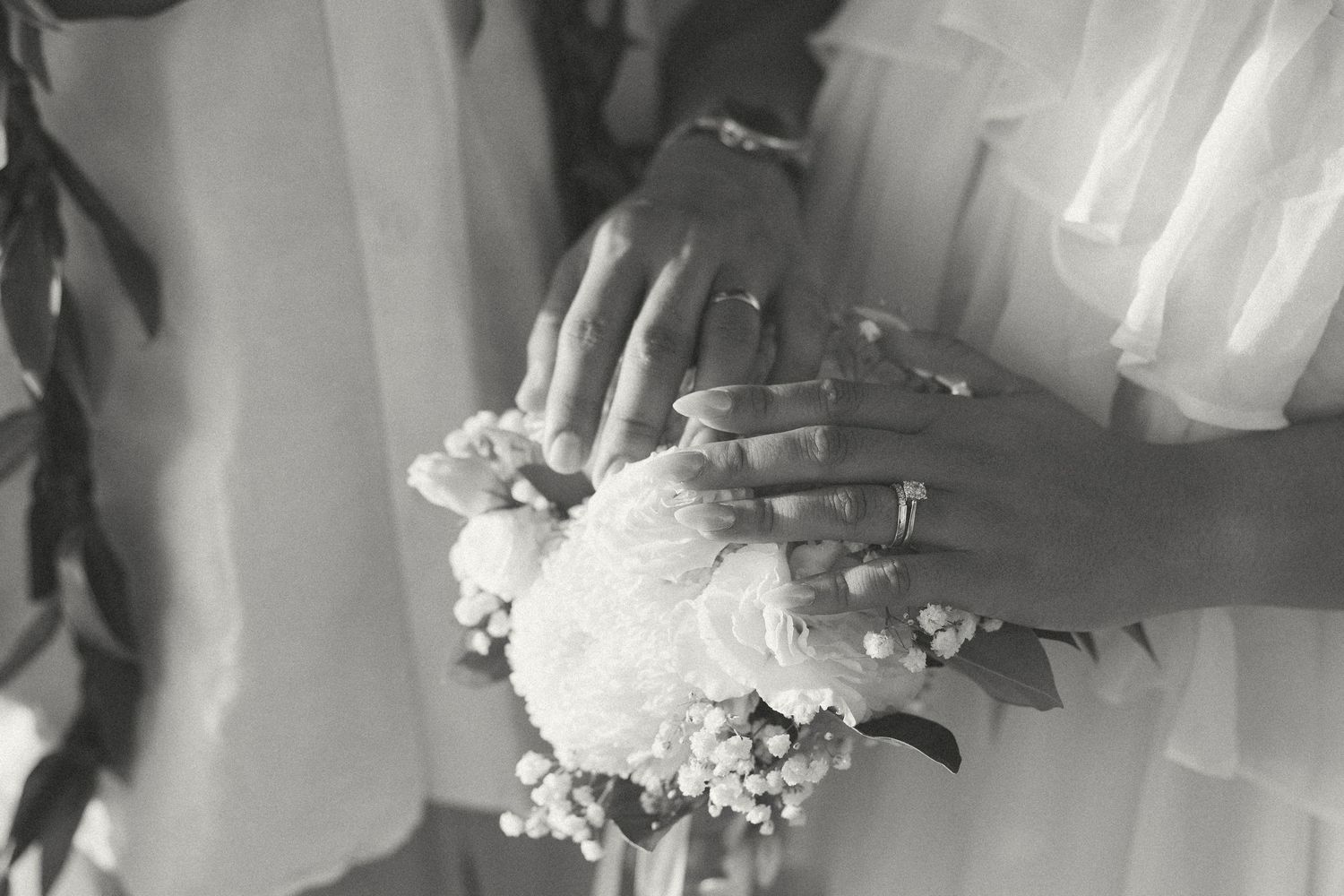Black and white photo of two hands with rings resting on a white bridal bouquet of roses and lily of the valley.