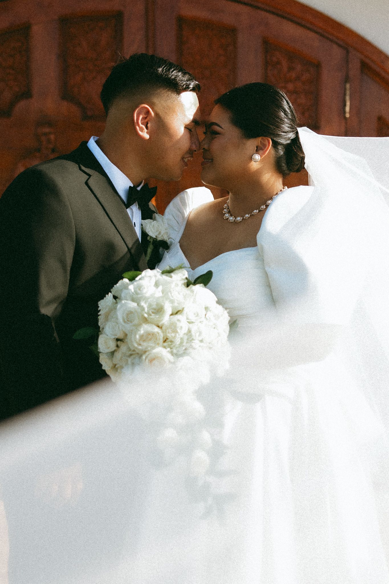 Bride and groom share a romantic kiss holding white rose bouquet, bride in elegant gown with flowing veil at church.