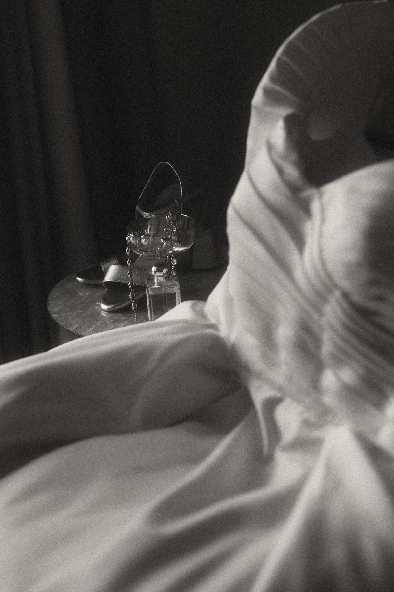 Black and white photo of a flowing wedding dress beside elegant bridal heels on a small table.