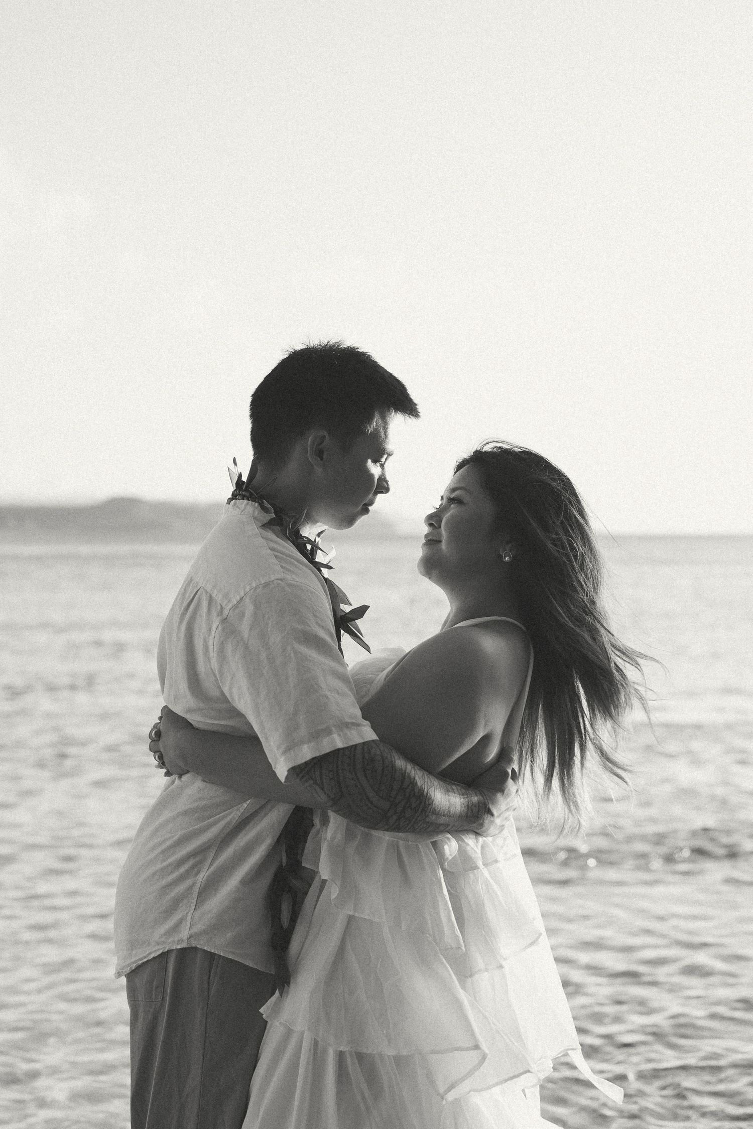 Black and white photo of a couple embracing on a beach, smiling at each other with ocean waves in the background.