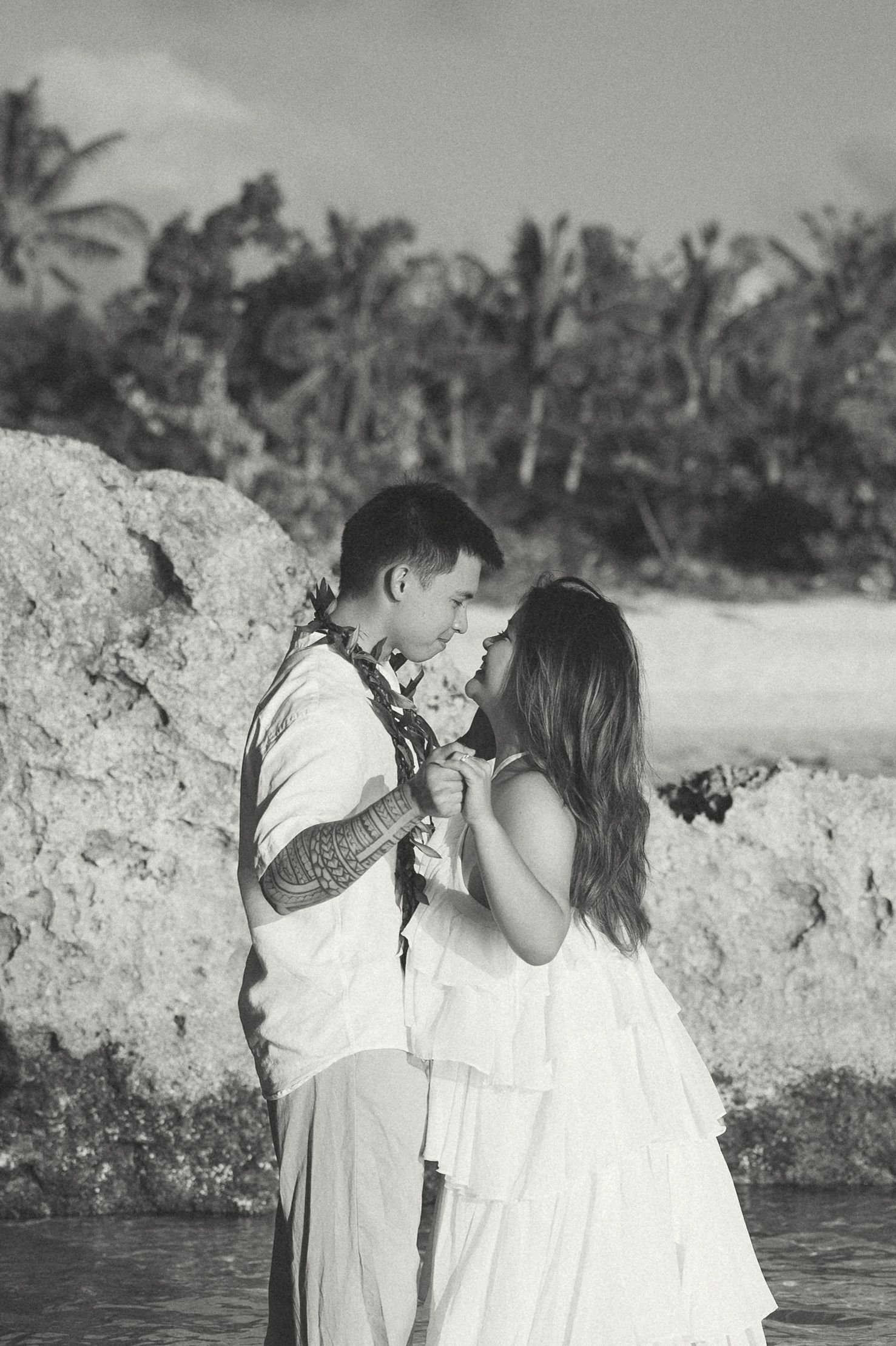 Black and white photo of a couple sharing a romantic moment on a tropical beach, with lush palm trees in the background.