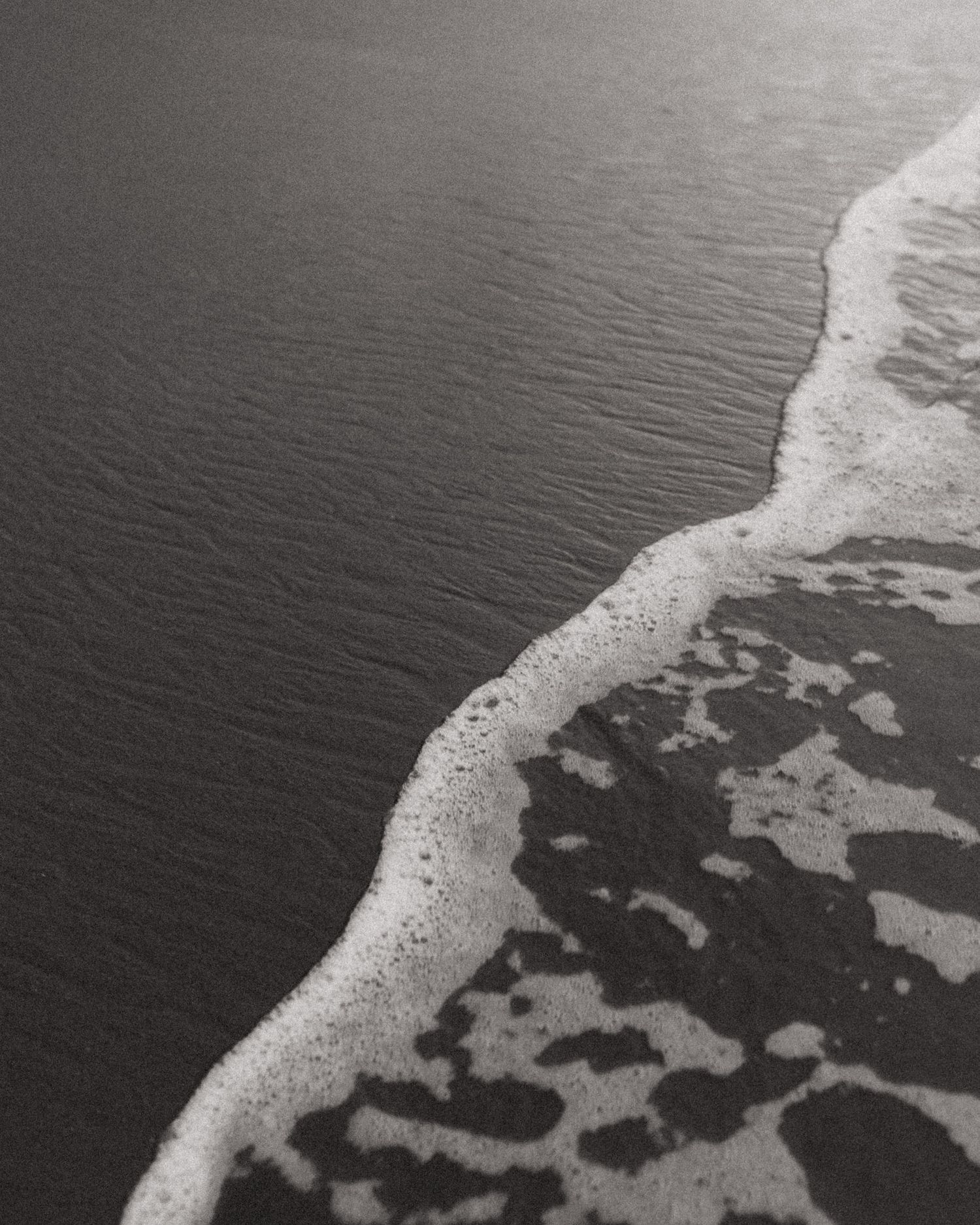 Black and white aerial view of ocean waves washing onto a sandy beach with foamy surf patterns.