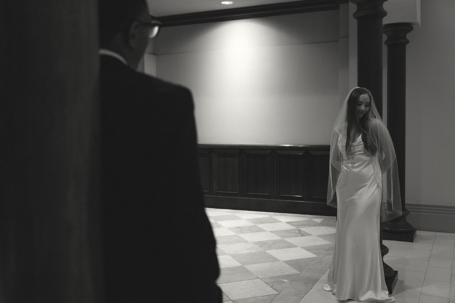 Black and white photo of a bride in a white gown and veil standing in an elegant hall, viewed by a groom in a suit.