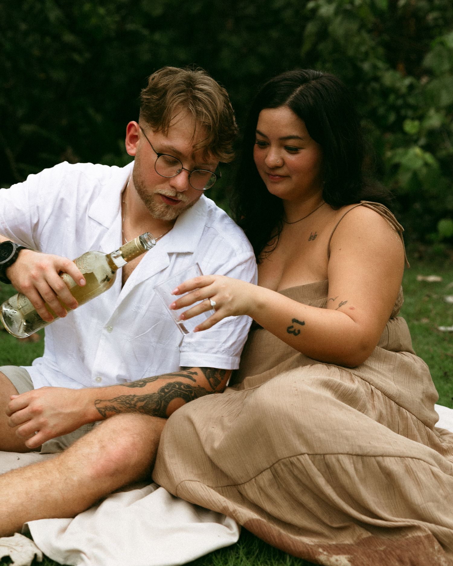 Couple sitting on grass sharing white wine at outdoor picnic, man pouring drink for woman in tan dress.