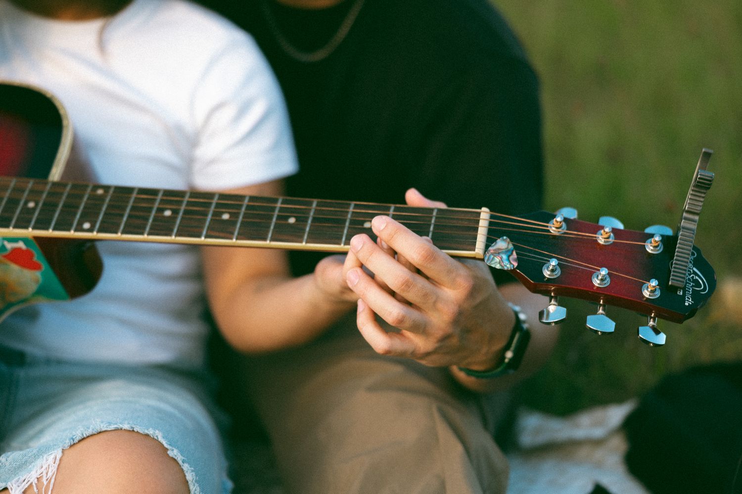 Two people sitting outdoors playing an acoustic guitar together on a sunny day.