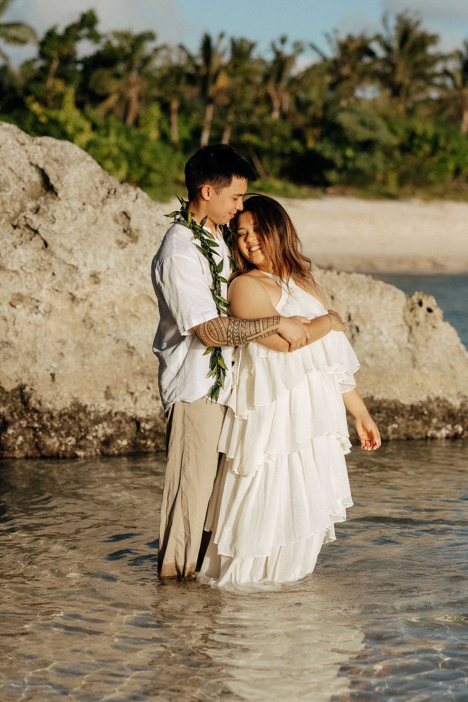 Couple embracing in shallow ocean water during beach wedding, groom wearing lei, bride in white ruffled dress, tropical backdrop.