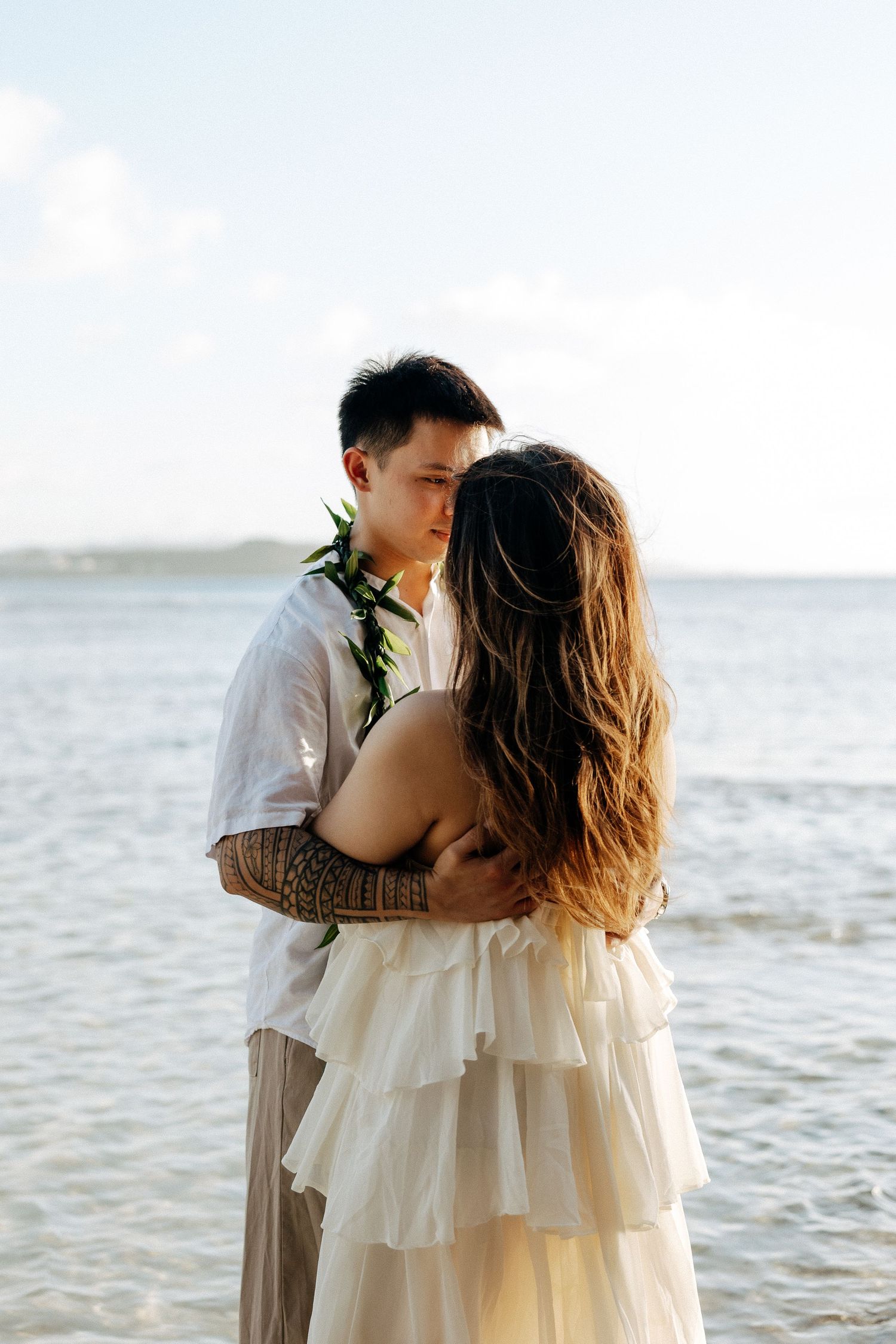 Couple embracing on a tropical beach, man wearing a floral lei and tattoos, woman in a white ruffled dress.