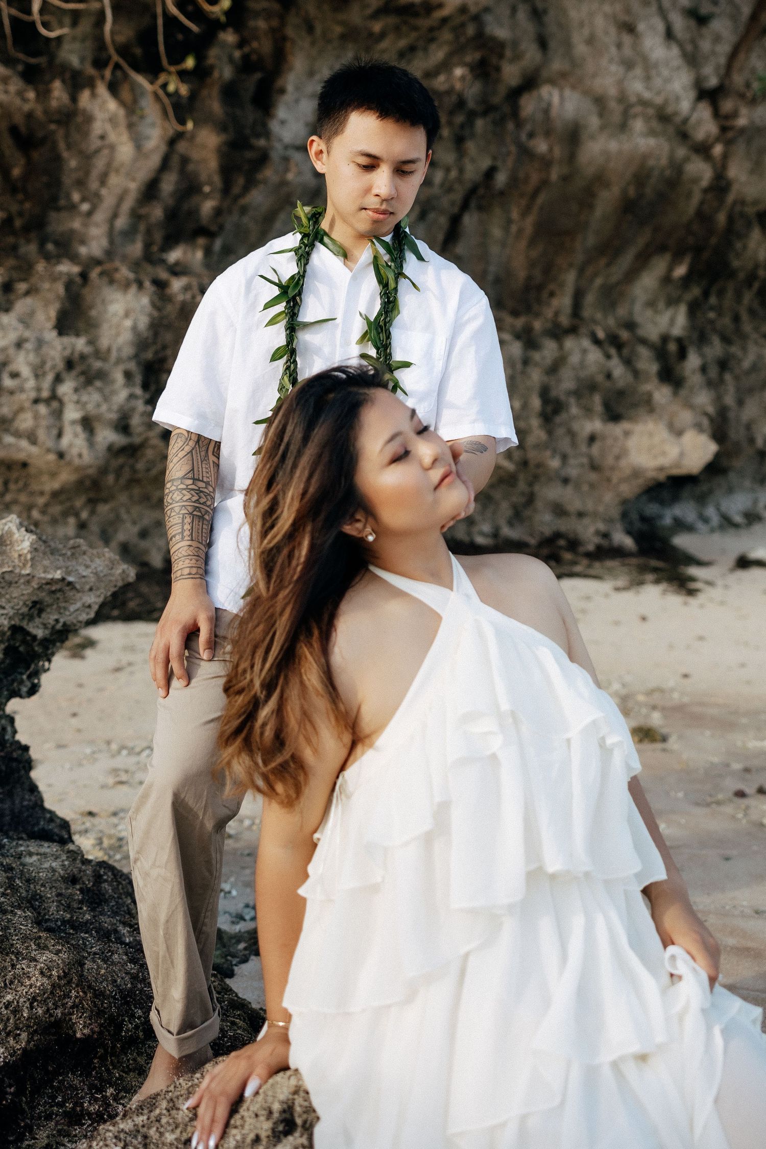 Couple in white attire poses on a rocky beach, man wearing a green lei and woman in a ruffled dress.