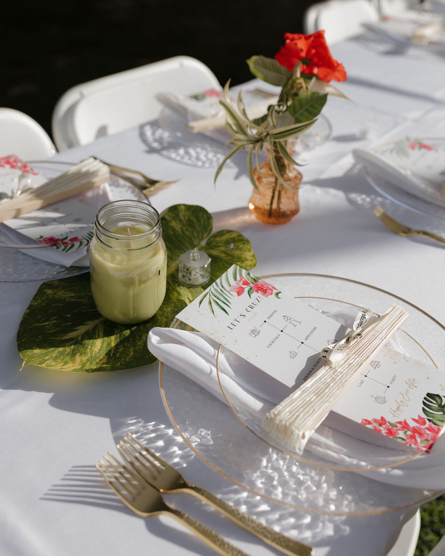 Elegant outdoor wedding table setting with gold cutlery, glass plates, green leaves, floral menu cards, and a red flower centerpiece.