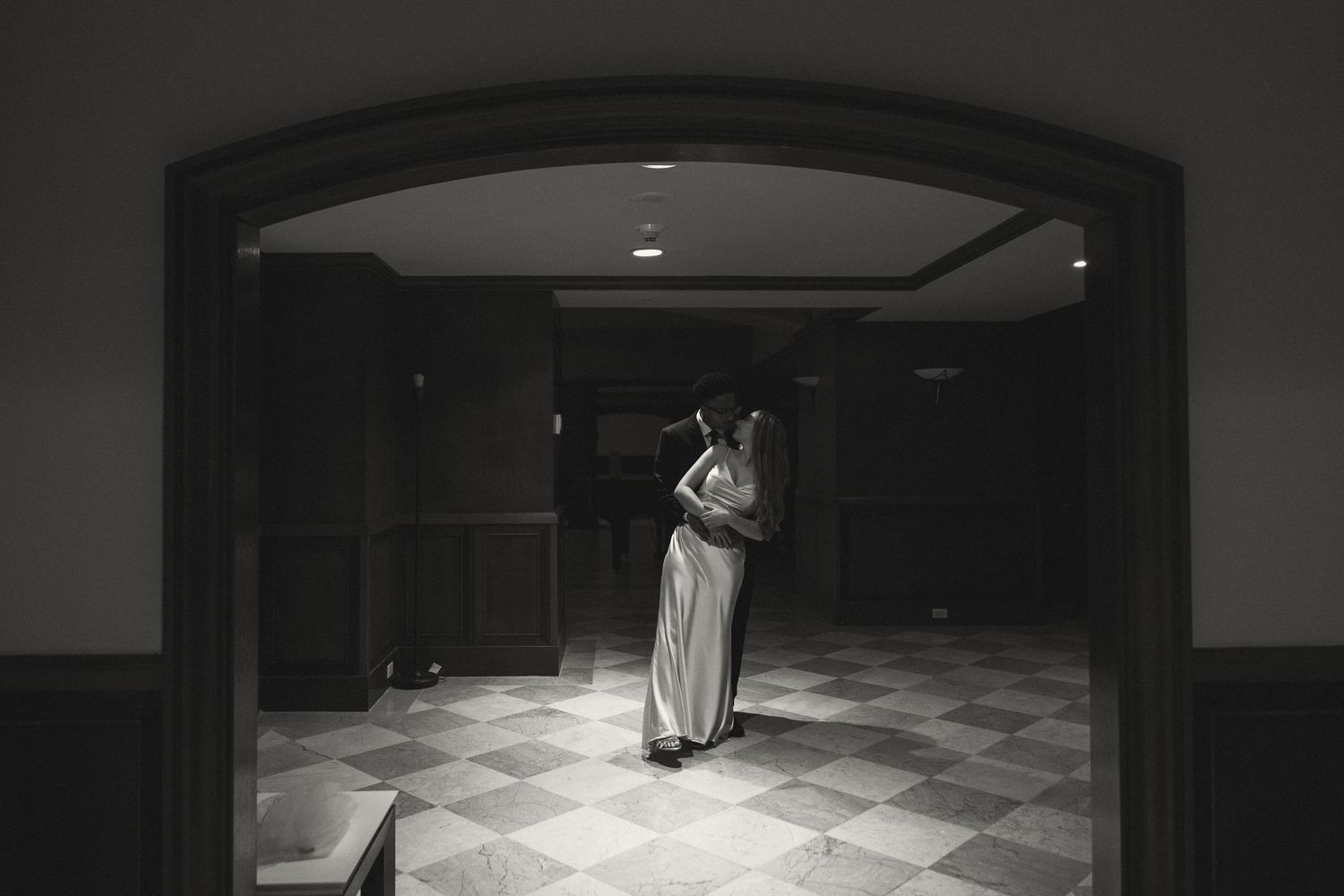 Black and white photo of a bride in a white gown standing in a dimly lit elegant hallway.