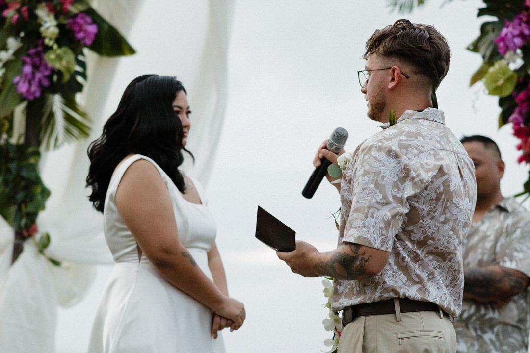 Couple exchanging wedding vows outdoors under floral arch, groom holding microphone in tropical setting.