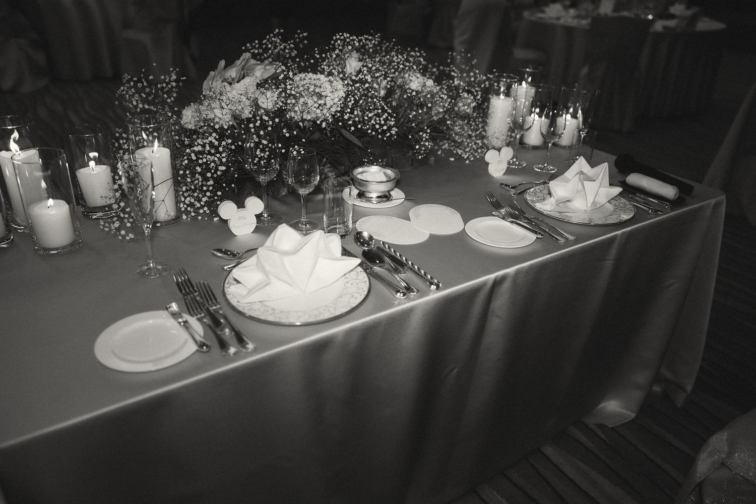 Black and white photo of an elegant wedding reception table set with candles, floral centerpiece, and neatly folded napkins.