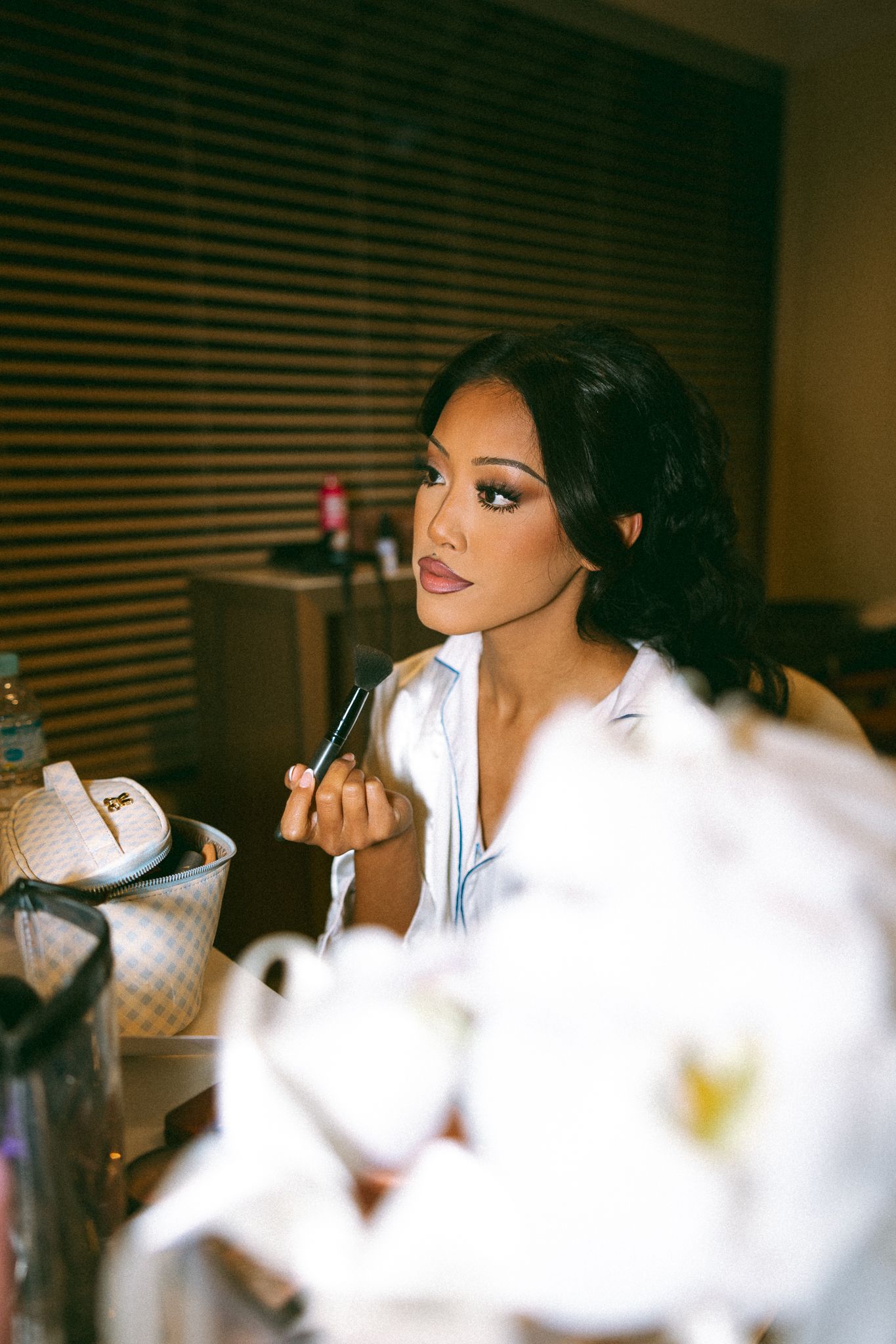 A beautiful bride applies makeup while seated at a vanity, wearing a white shirt with flowers in the foreground.