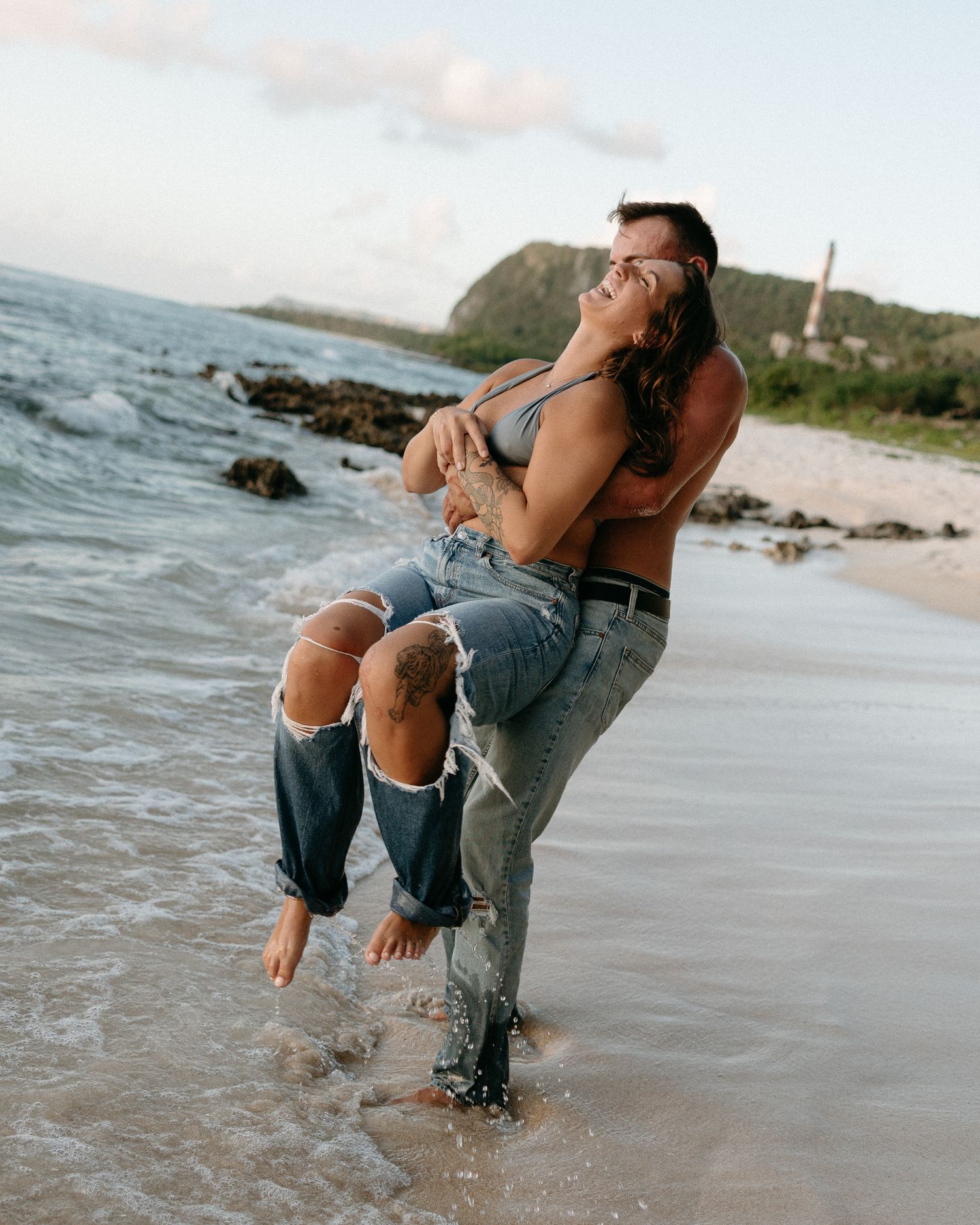 Couple embracing on a tropical beach, man lifting woman in ripped jeans, waves splashing, green hills in background.