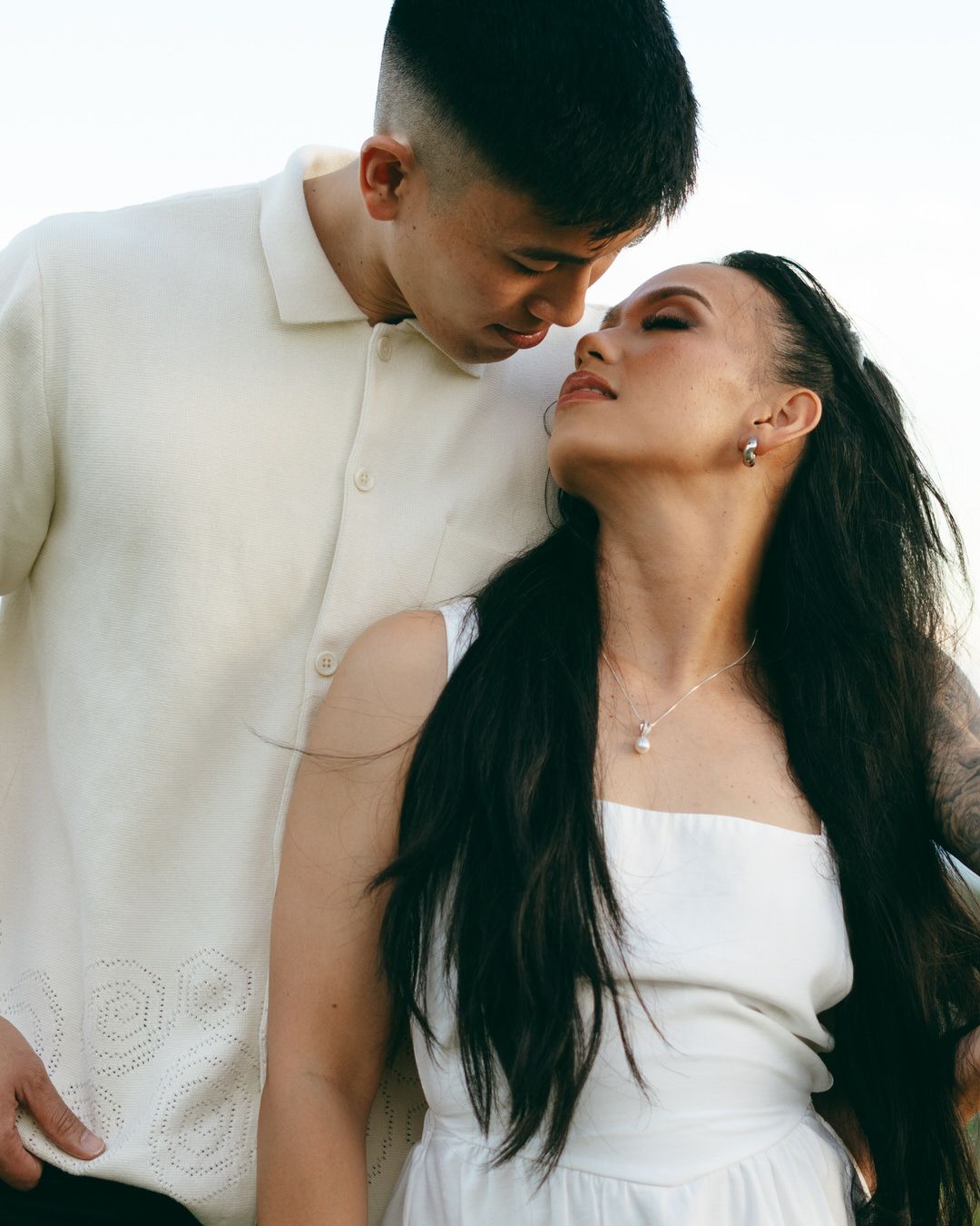A couple in white outfits share a romantic moment, the man leaning toward the woman with long black hair outdoors.