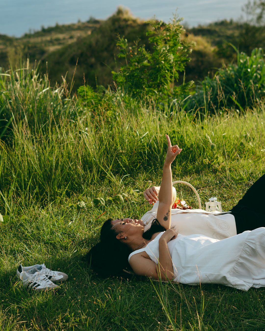 A woman lies on green grass at a picnic, pointing upward and smiling, with a wicker basket nearby.
