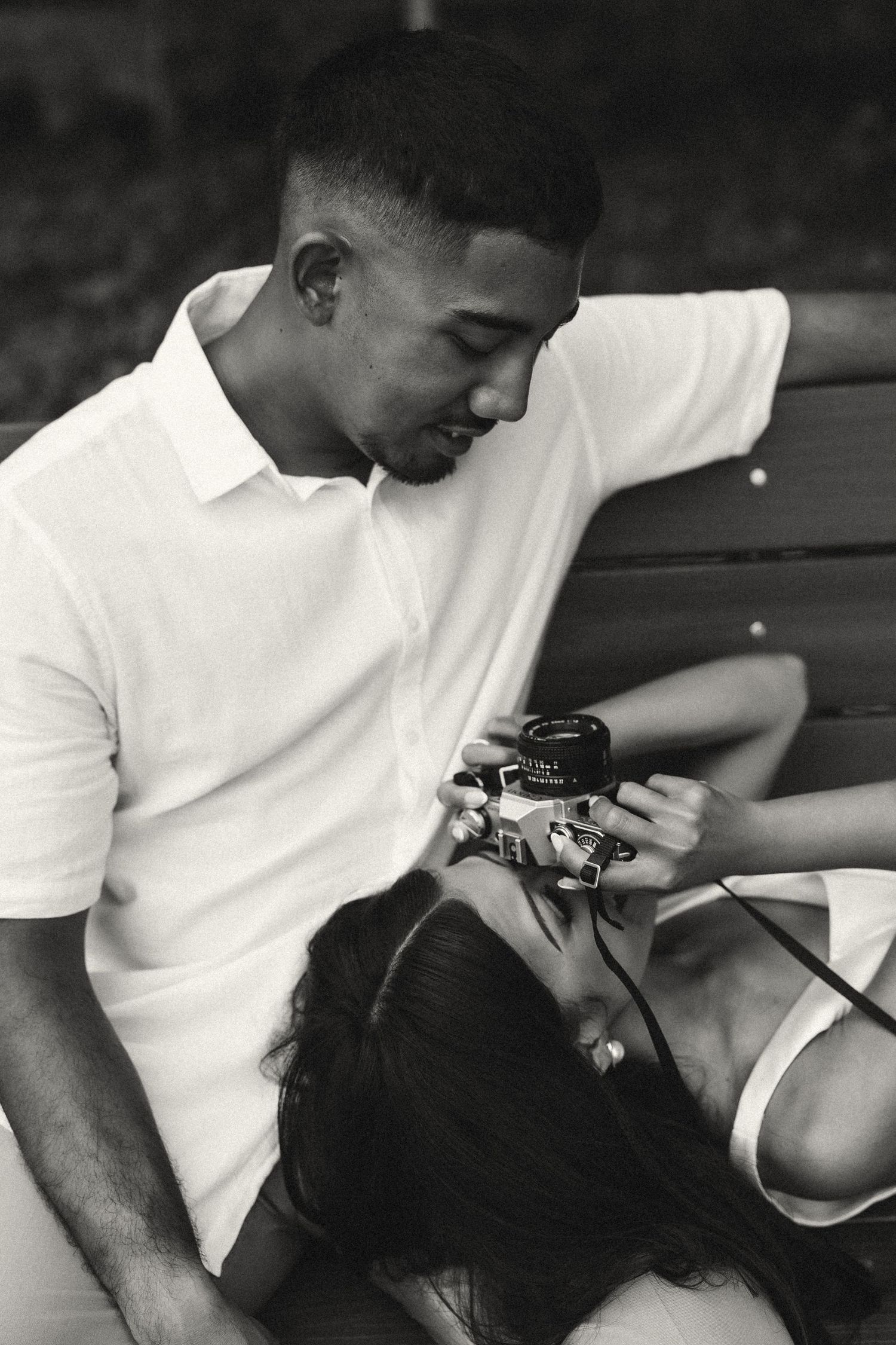 Black and white photo of a young couple sitting on a bench, looking at a vintage film camera together.