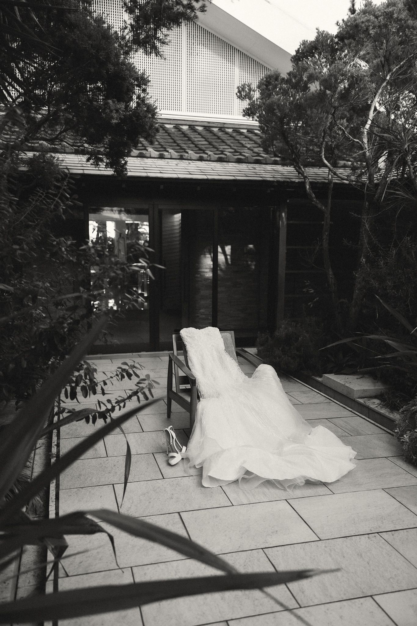 Black and white photo of a white wedding dress draped over a chair on a stone patio outside a modern home.