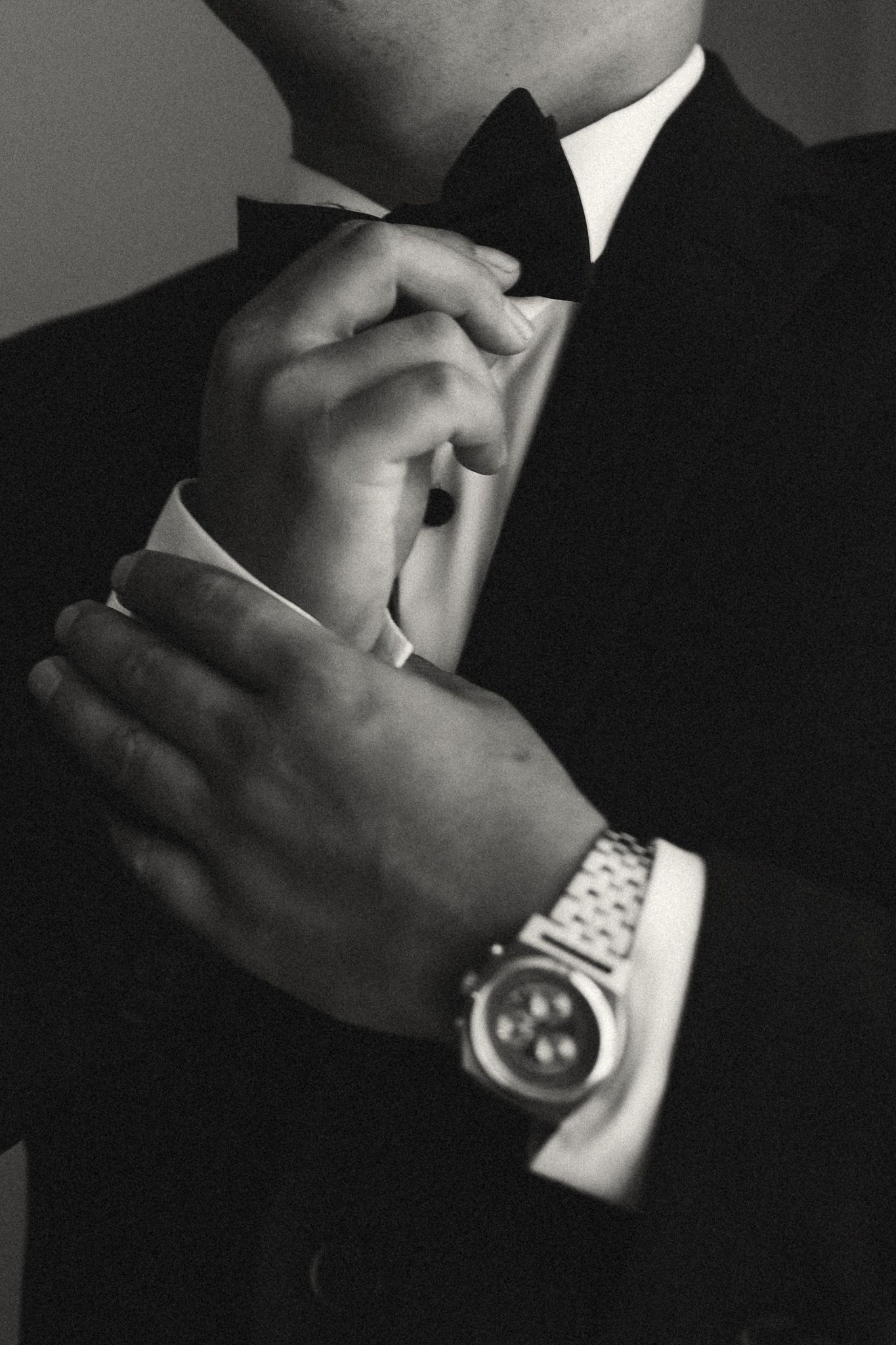 Man in tuxedo adjusting bow tie while wearing a luxury watch, black and white formal elegance photo.