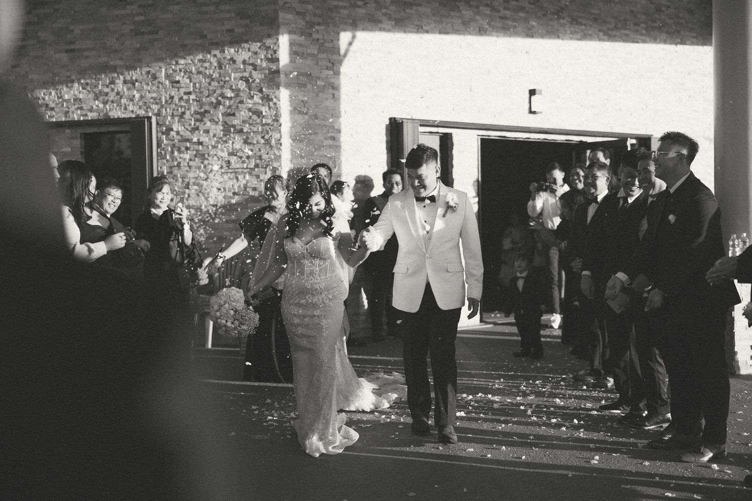 Black and white photo of a bride and groom exiting their wedding venue surrounded by cheering guests.