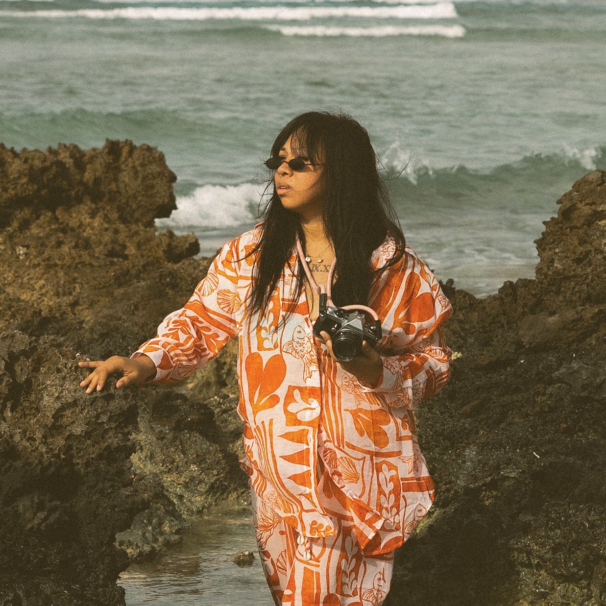 Woman in orange printed outfit holding camera poses on rocky shoreline with ocean waves in background.