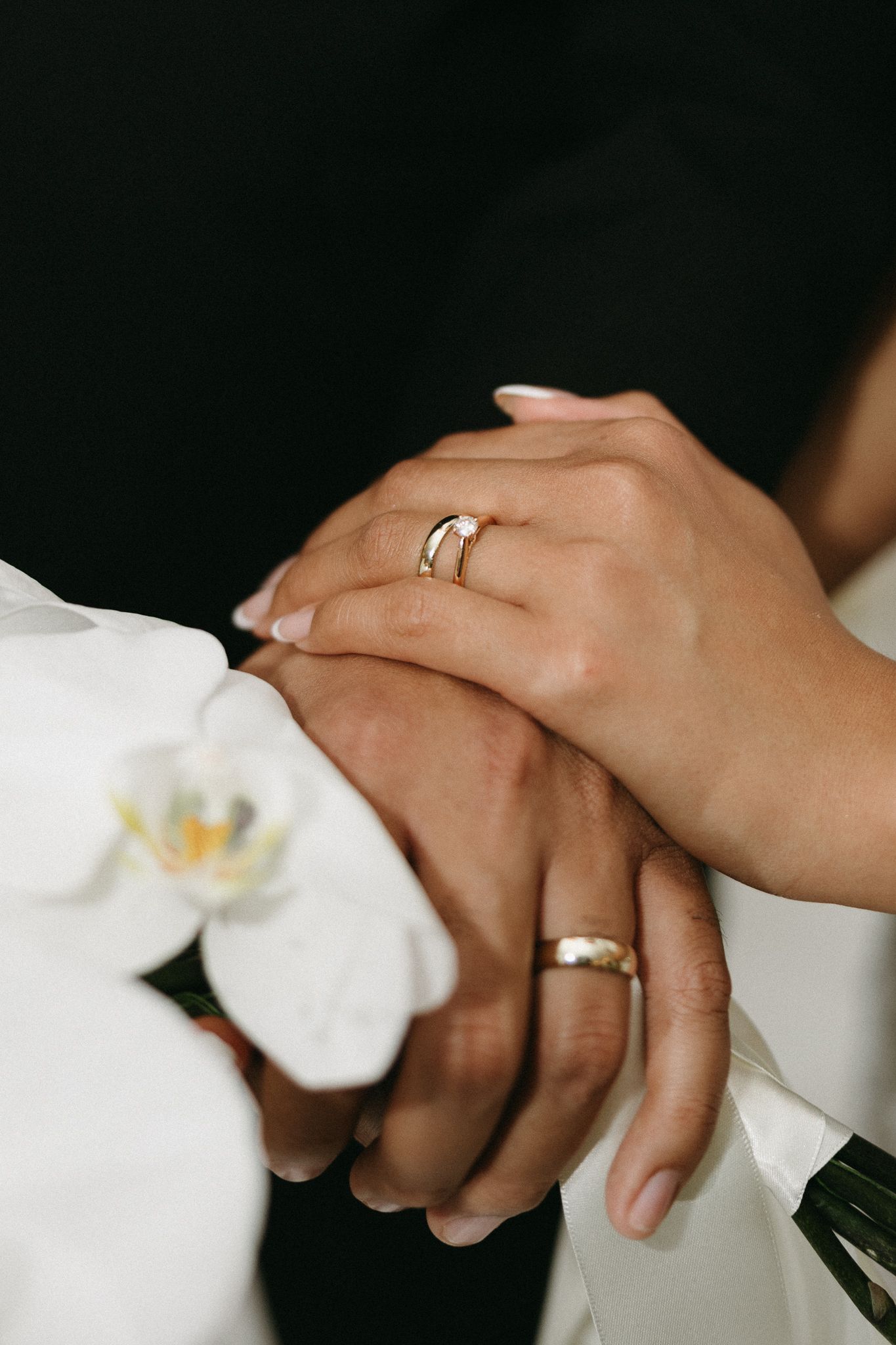 Bride and groom holding hands showing wedding rings, with white orchid flowers in foreground.