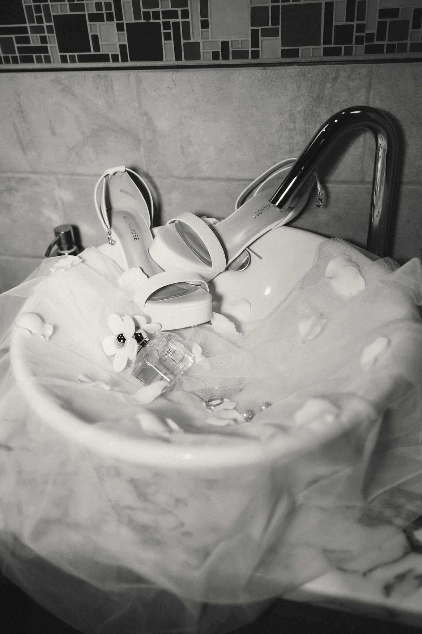 Black and white photo of bridal heels and perfume resting in a white bathroom sink with tulle veil.