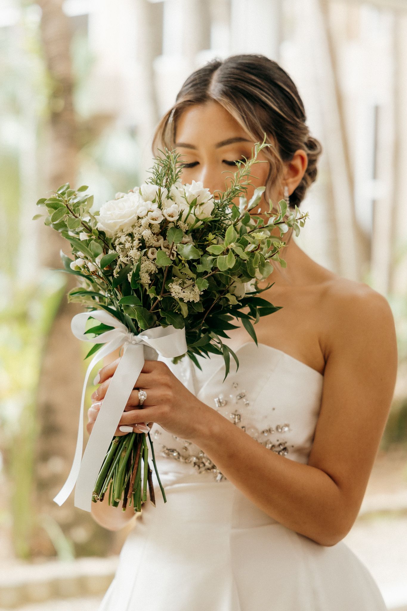 Bride in strapless white gown smelling a lush white and green wedding bouquet tied with ribbon.