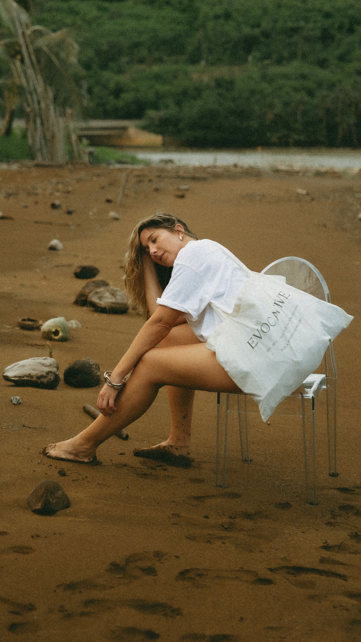 Woman in white dress sitting on clear chair on sandy beach holding Evocame bag with green trees in background.