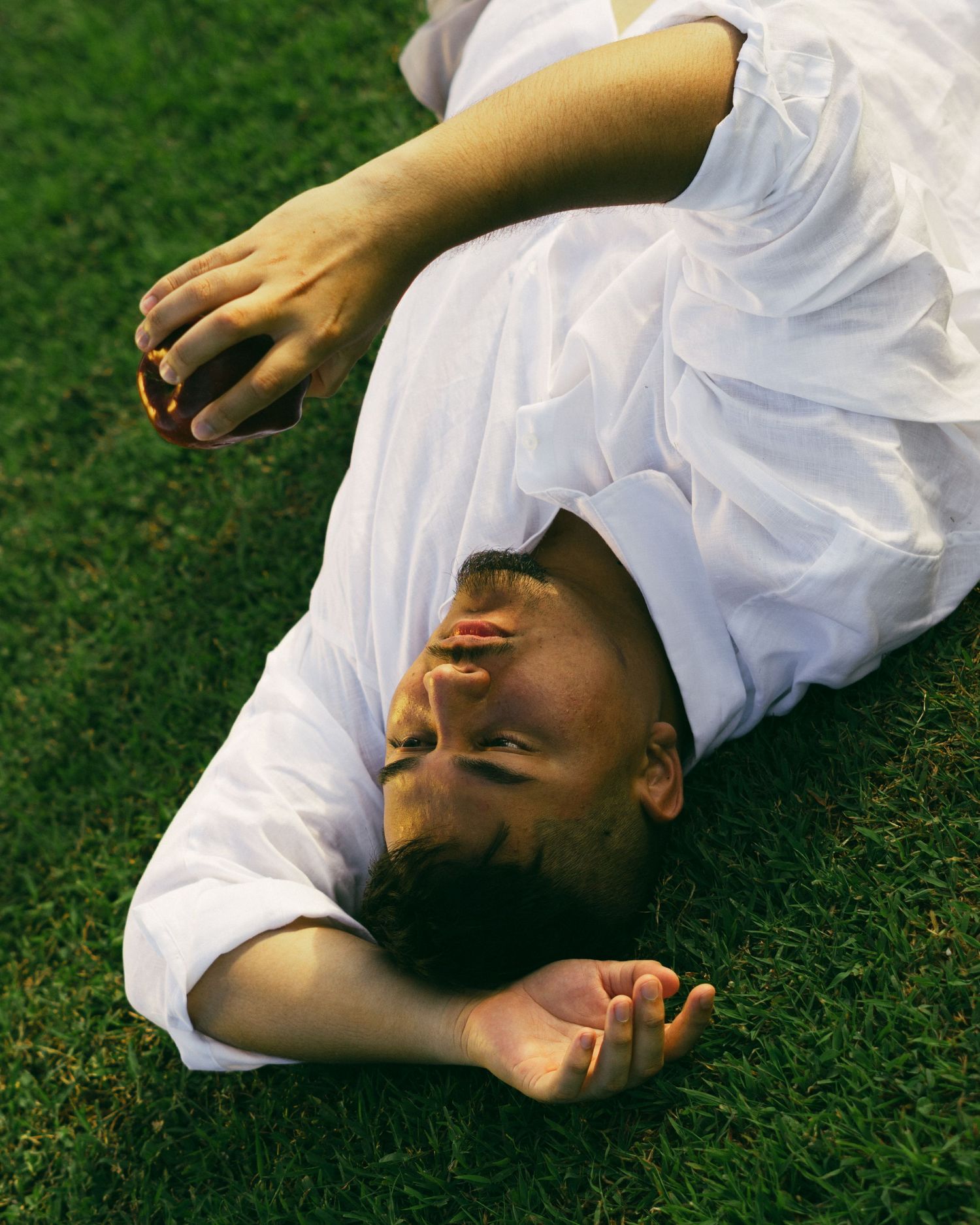 Person in white dress lying on green grass, looking upward with a relaxed expression.