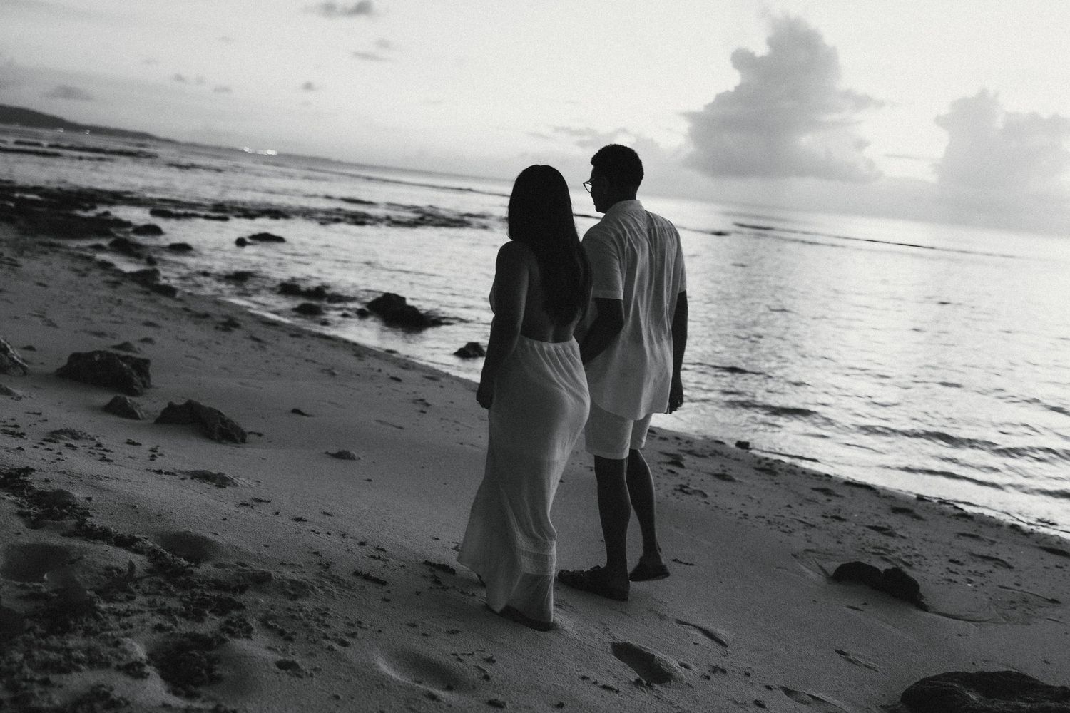Black and white photo of a couple walking hand in hand on a sandy beach at sunset near the ocean.