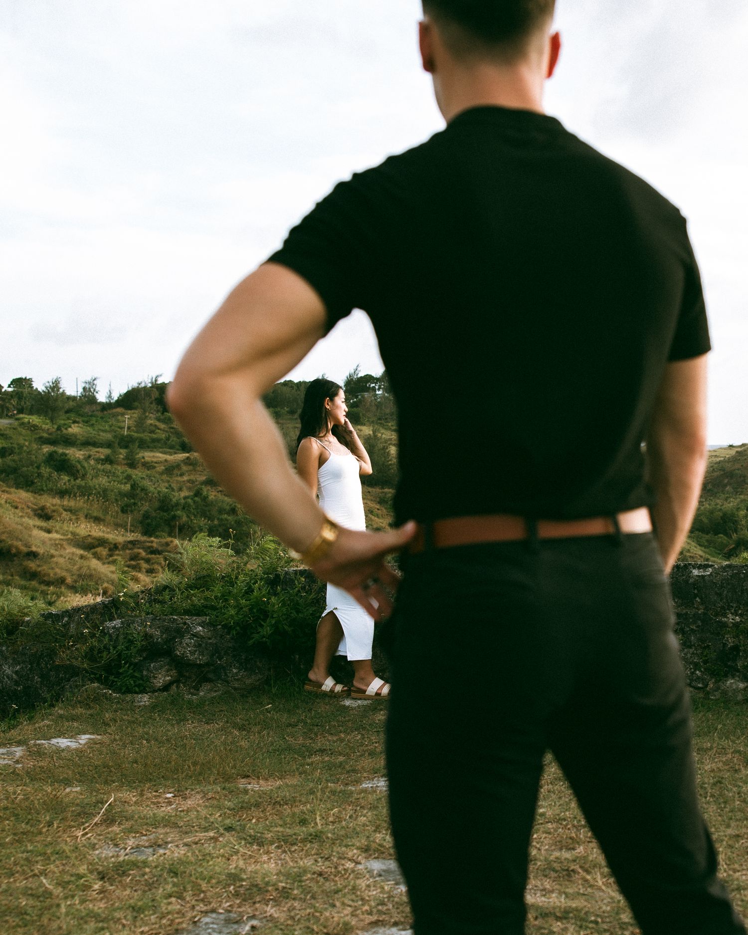 Man in black shirt and pants stands in field facing woman in white dress on a green hillside.