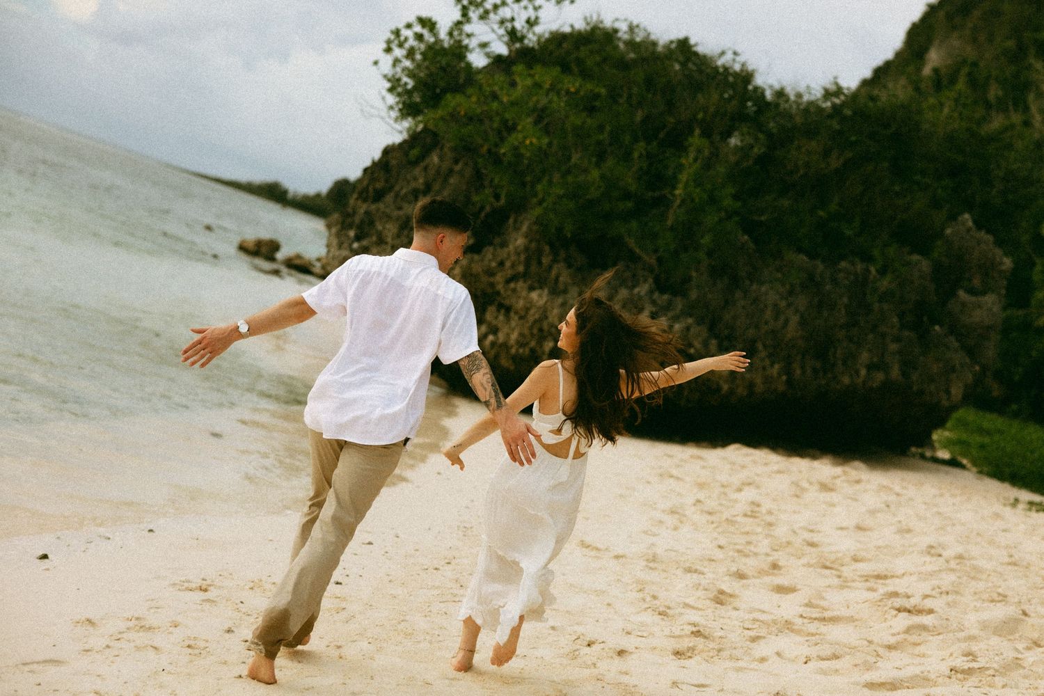 Couple dancing barefoot on a tropical sandy beach near rocky cliffs during a romantic outdoor photoshoot.
